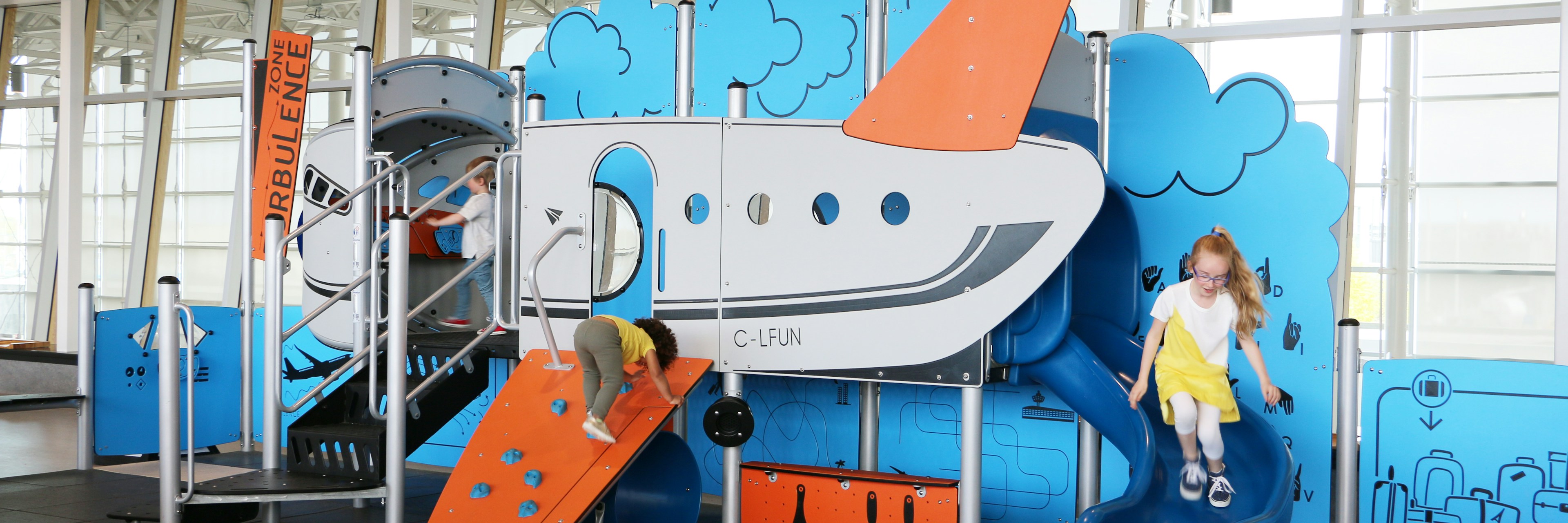 Deux enfants jouent dans une aire de jeux couverte sur le thème de l'avion, équipée d'un toboggan et d'un mur d'escalade. Des couleurs vives, bleu et orange, ornent la structure de jeux.