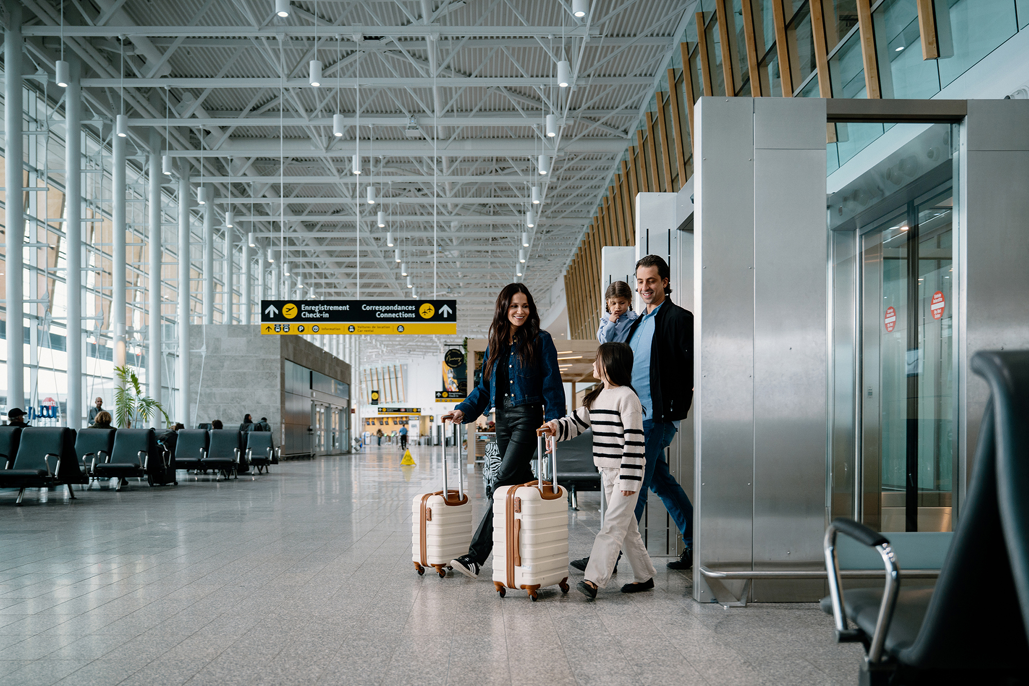Une famille de quatre personnes, accompagnée de ses bagages, traverse un terminal d'aéroport spacieux et moderne. On aperçoit des panneaux indicateurs et des coins où s'asseoir ; la famille semble heureuse et prête à partir.