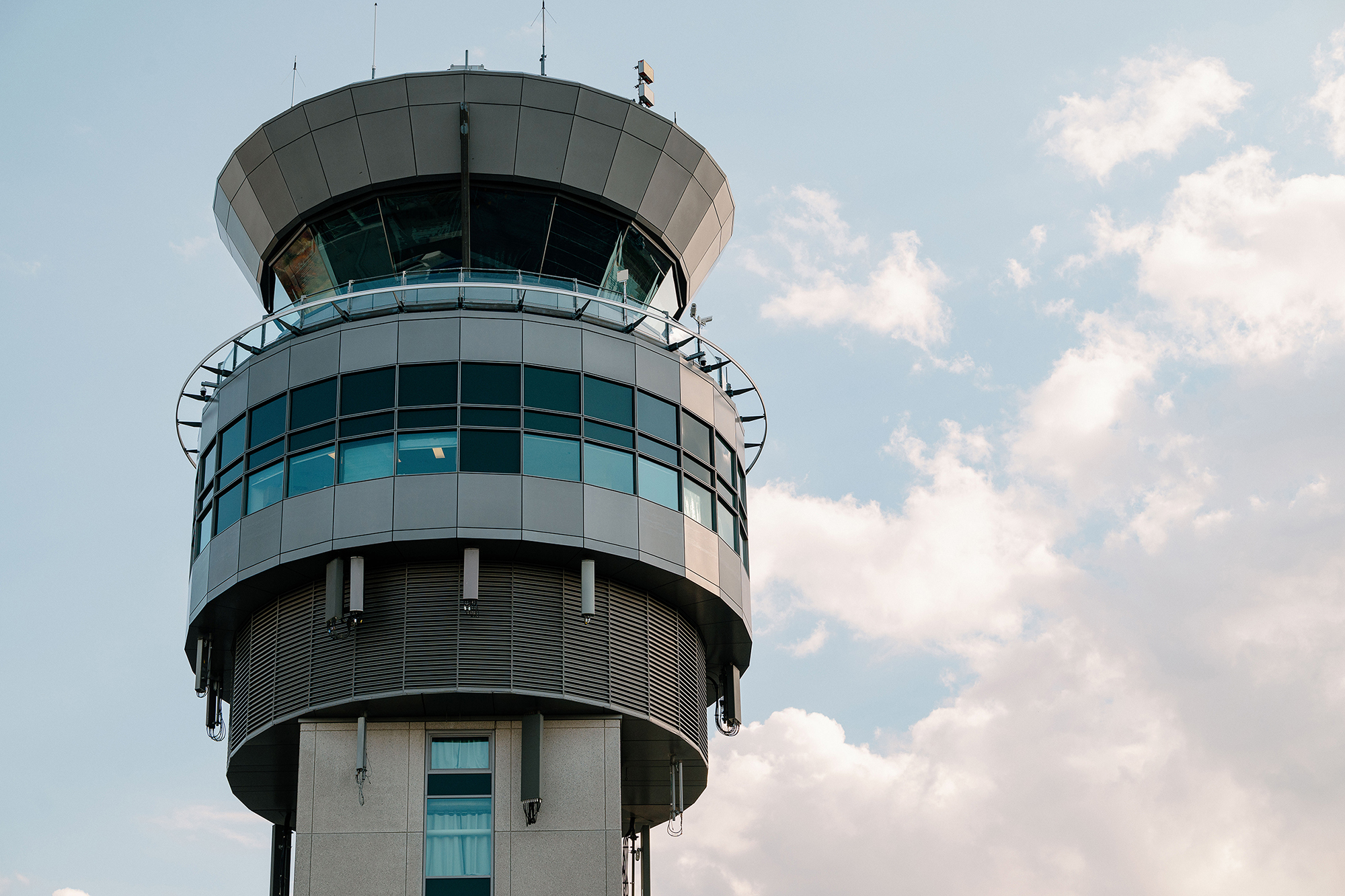 A modern control tower with large windows stands out against a partly cloudy sky; viewed from below, sunlight reflects off the glass.