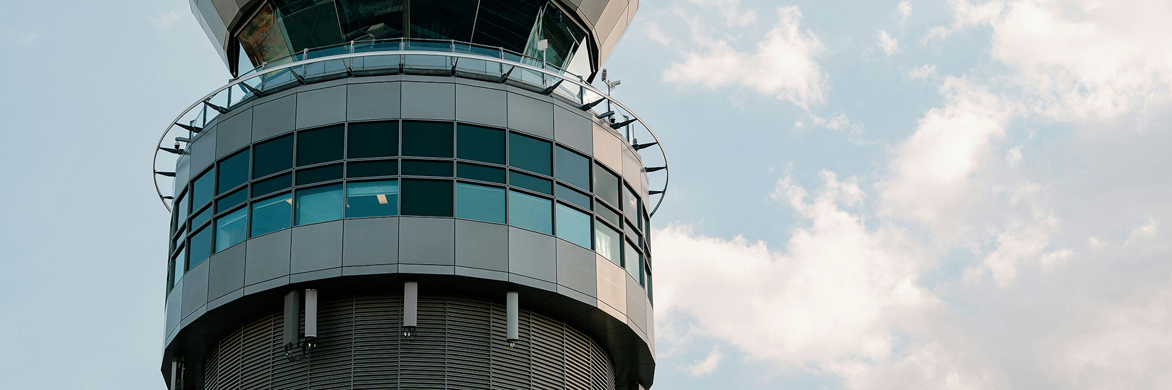 A modern control tower with large windows stands out against a partly cloudy sky; viewed from below, sunlight reflects off the glass.