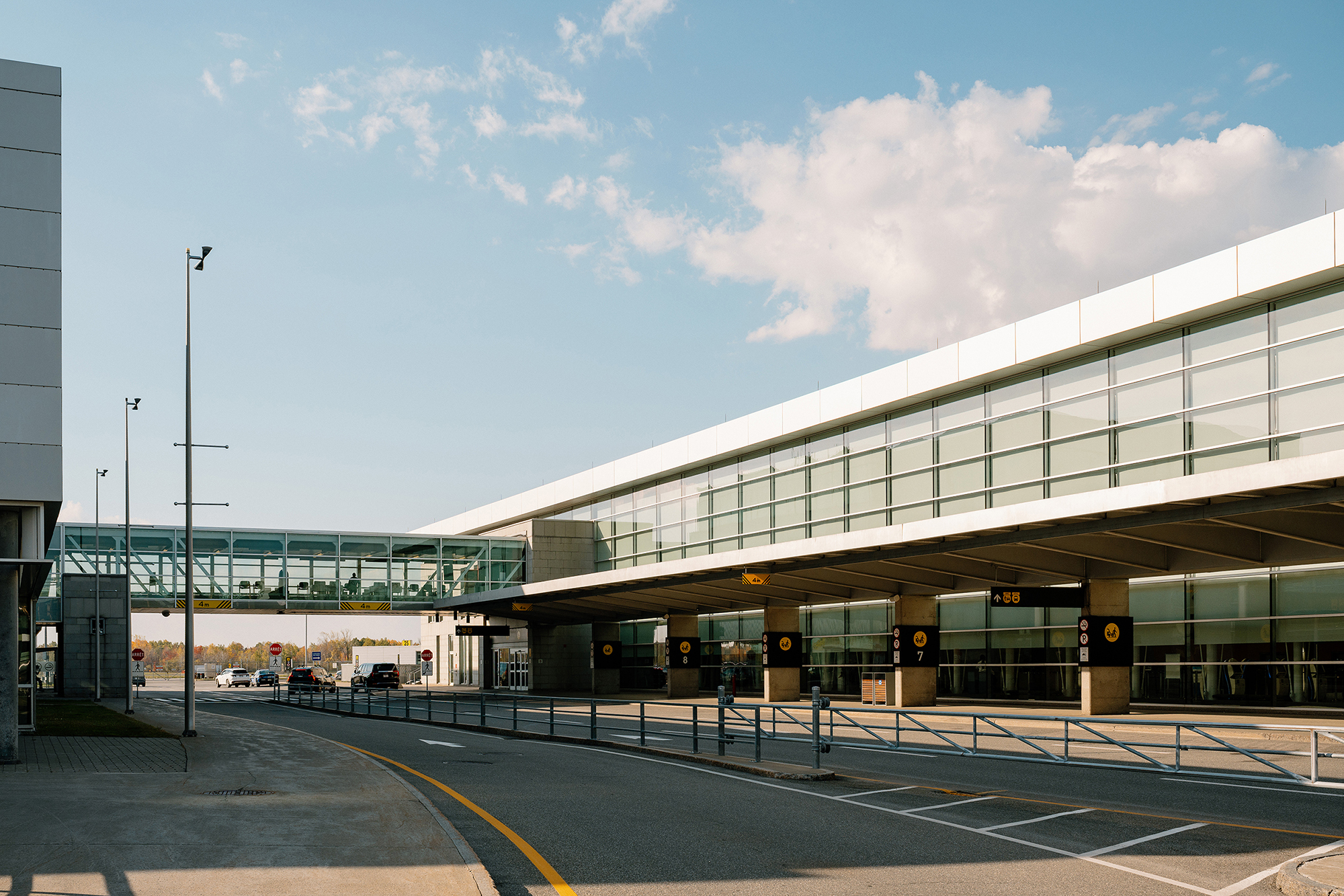 Un terminal d'aéroport moderne doté de grandes baies vitrées, d'une passerelle reliant les bâtiments et d'une voie de circulation presque déserte, sous un ciel bleu limpide parsemé de quelques nuages.