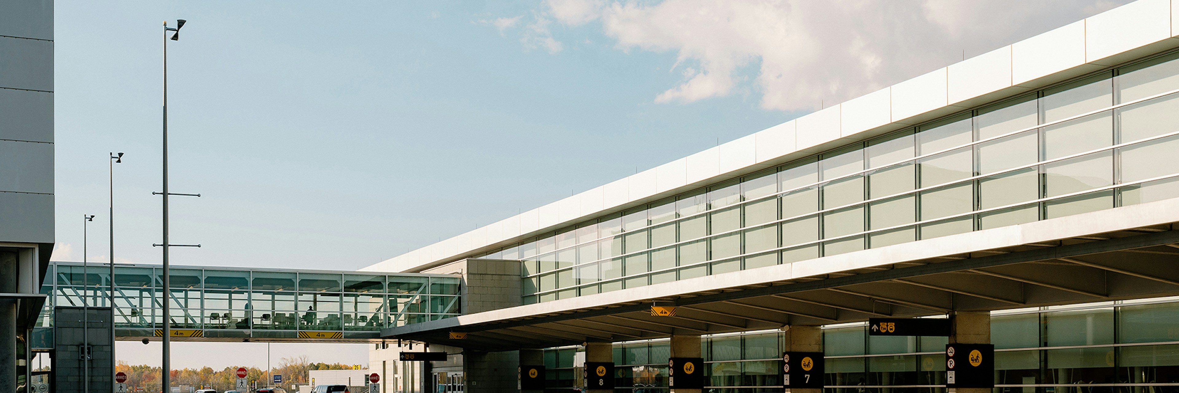 Un terminal d'aéroport moderne doté de grandes baies vitrées, d'une passerelle reliant les bâtiments et d'une voie de circulation presque déserte, sous un ciel bleu limpide parsemé de quelques nuages.