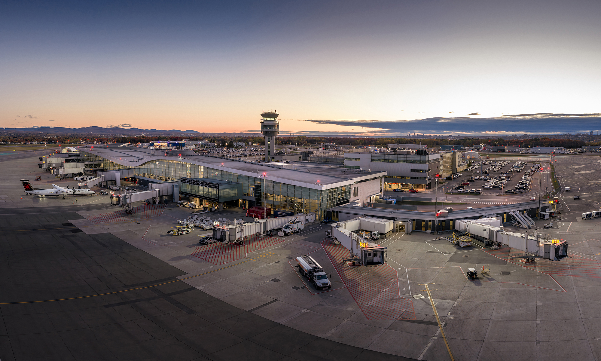 Un terminal d'aéroport au coucher du soleil, avec plusieurs avions stationnés aux portes d'embarquement, des passerelles d'embarquement raccordées, des véhicules aéroportuaires à proximité, une tour de contrôle en arrière-plan et des rangées de voitures garées.