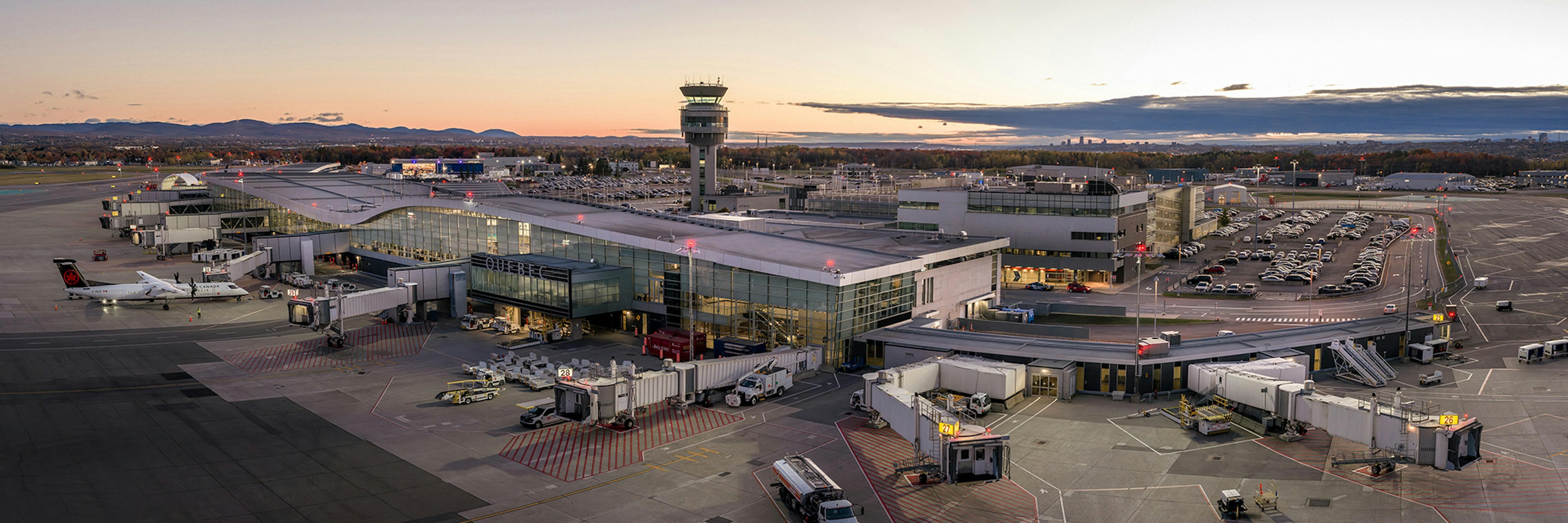 Un terminal d'aéroport au coucher du soleil, avec plusieurs avions stationnés aux portes d'embarquement, des passerelles d'embarquement raccordées, des véhicules aéroportuaires à proximité, une tour de contrôle en arrière-plan et des rangées de voitures garées.