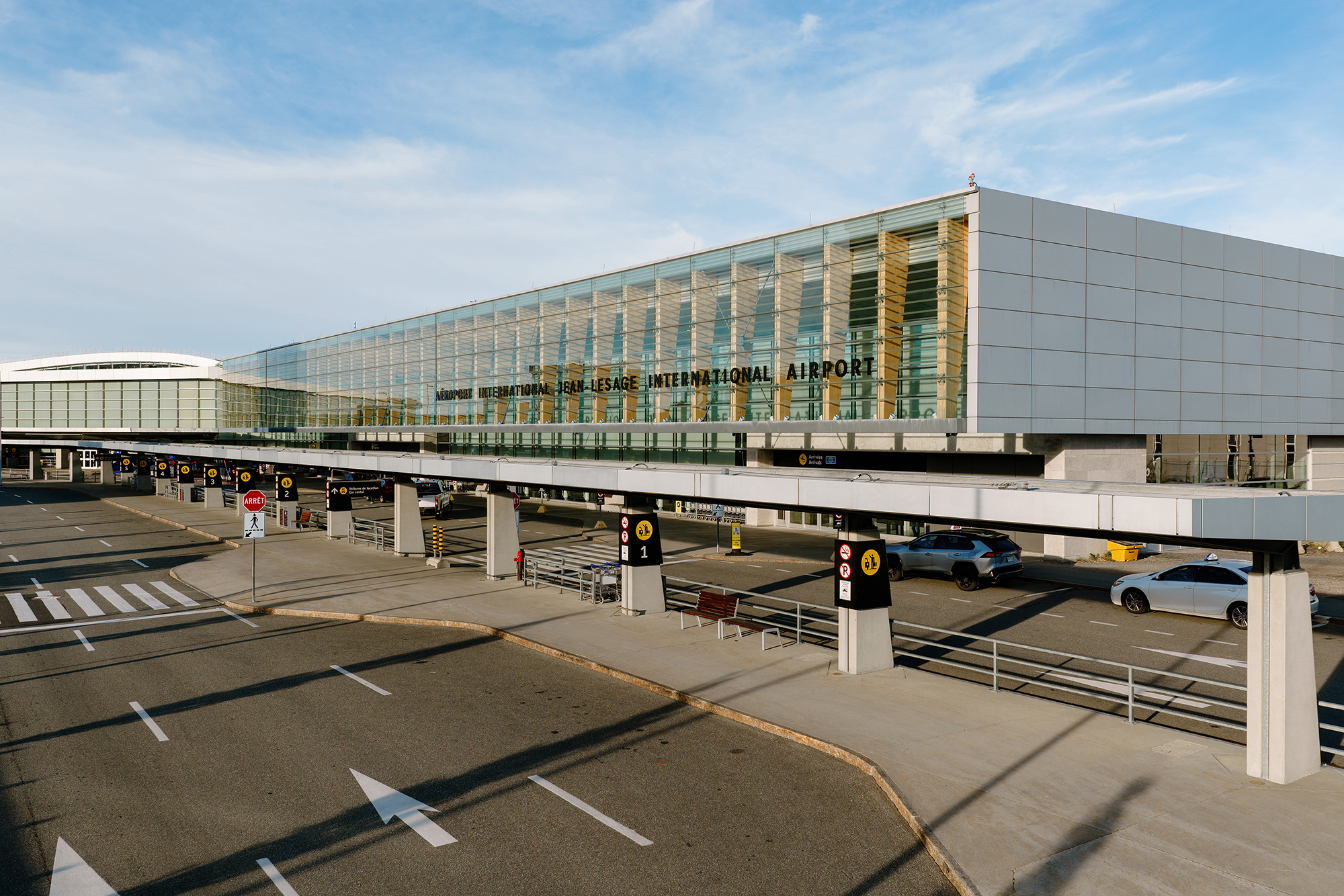 Extérieur d'un terminal d'aéroport moderne avec des baies vitrées, une enseigne bien visible indiquant « Aéroport international Ted Stevens », des voitures garées et circulant devant, et un ciel bleu limpide au-dessus.