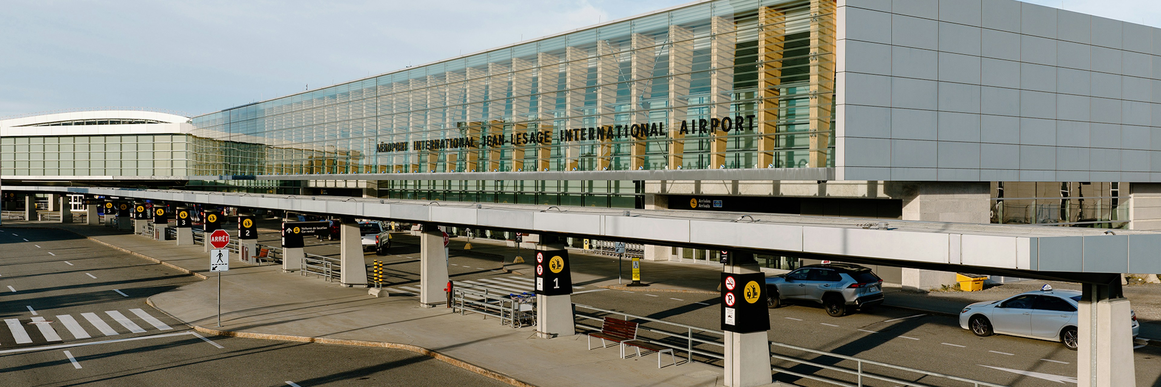 Extérieur d'un terminal d'aéroport moderne avec des baies vitrées, une enseigne bien visible indiquant « Aéroport international Ted Stevens », des voitures garées et circulant devant, et un ciel bleu limpide au-dessus.