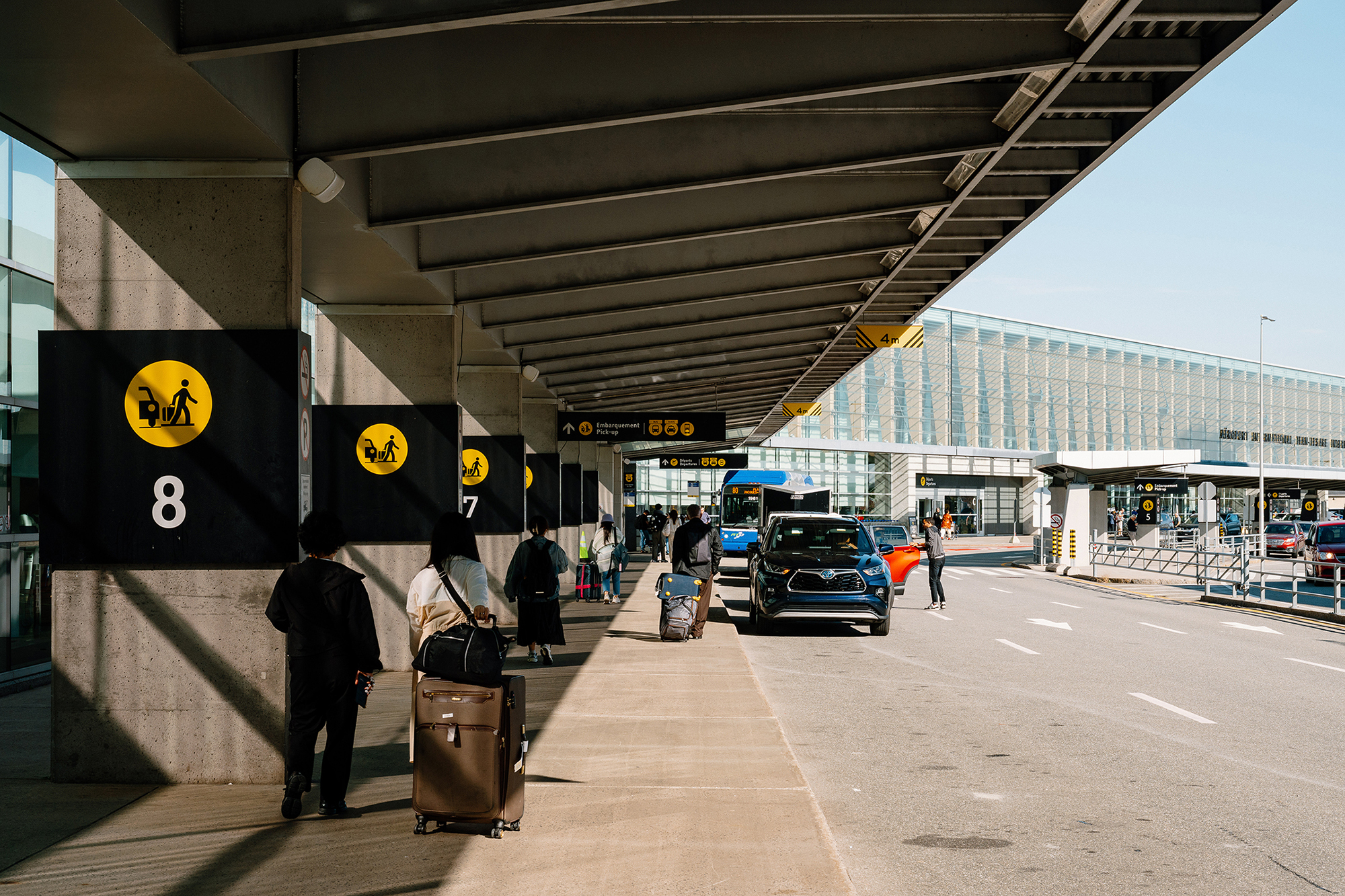 Des voyageurs chargés de bagages marchent sur un trottoir couvert à l'extérieur d'un terminal d'aéroport. Des voitures sont garées le long du trottoir, et des panneaux numérotés, suspendus aux colonnes, indiquent les zones de prise en charge. Le temps est clair et ensoleillé.