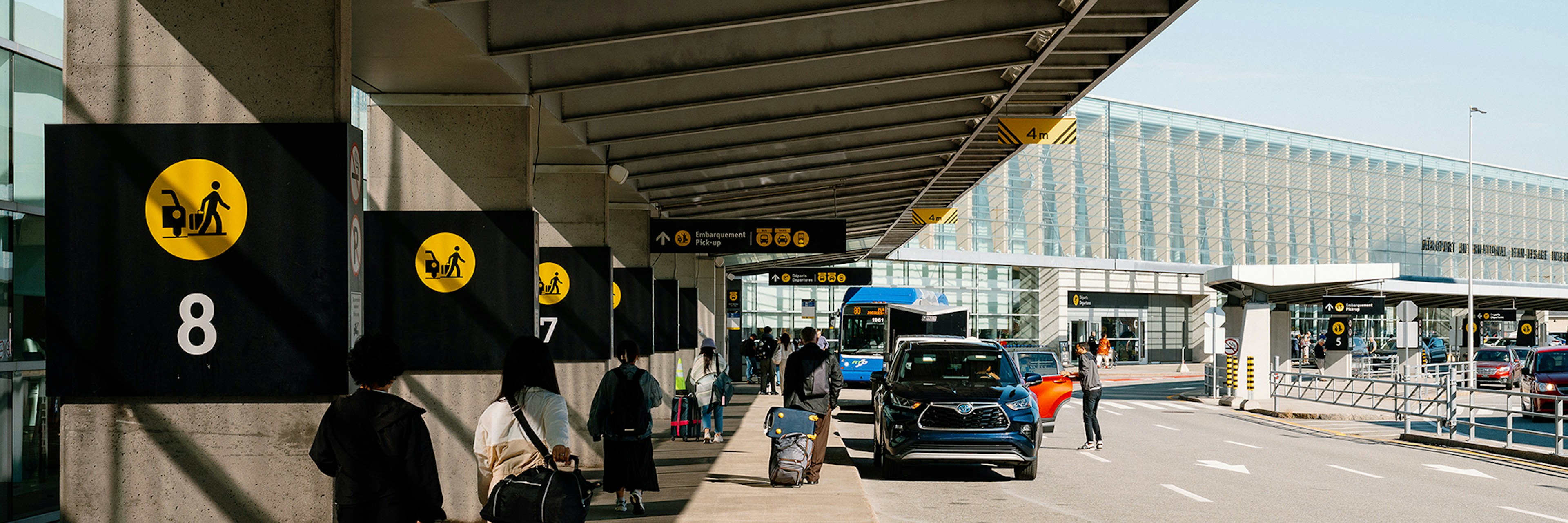 Des voyageurs chargés de bagages marchent sur un trottoir couvert à l'extérieur d'un terminal d'aéroport. Des voitures sont garées le long du trottoir, et des panneaux numérotés, suspendus aux colonnes, indiquent les zones de prise en charge. Le temps est clair et ensoleillé.