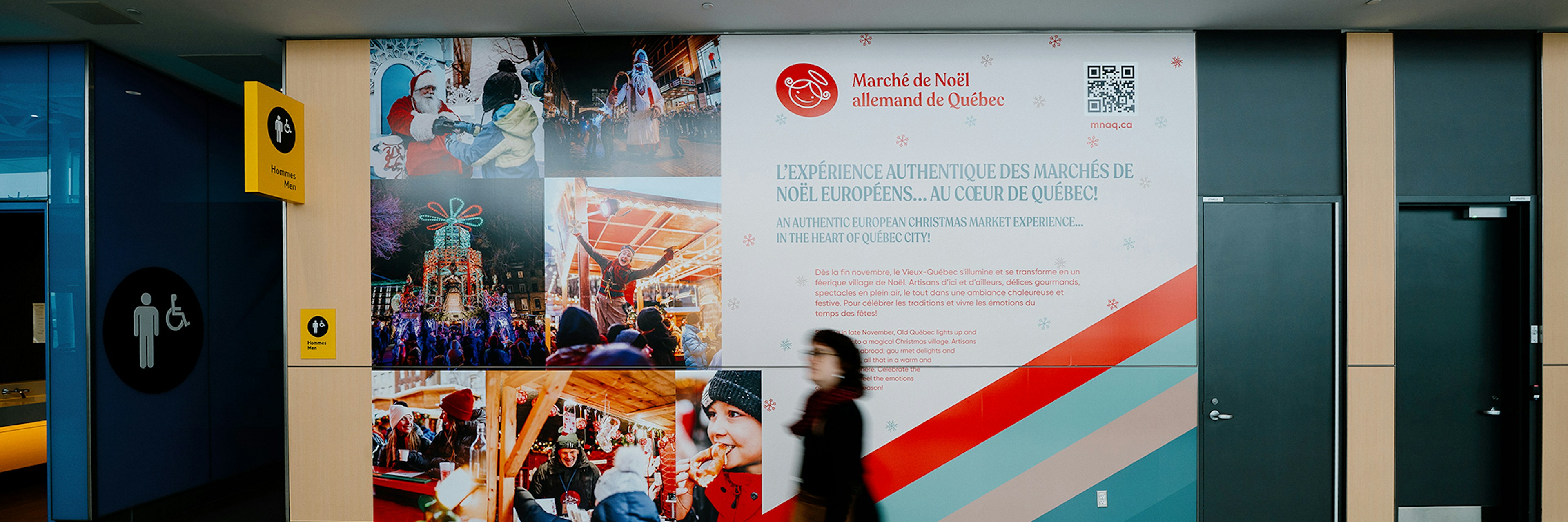 Une personne passe devant un grand panneau mural coloré présentant le marché de Noël allemand de Québec, situé dans un espace public couvert où l'on aperçoit, à gauche, des panneaux indiquant les toilettes.