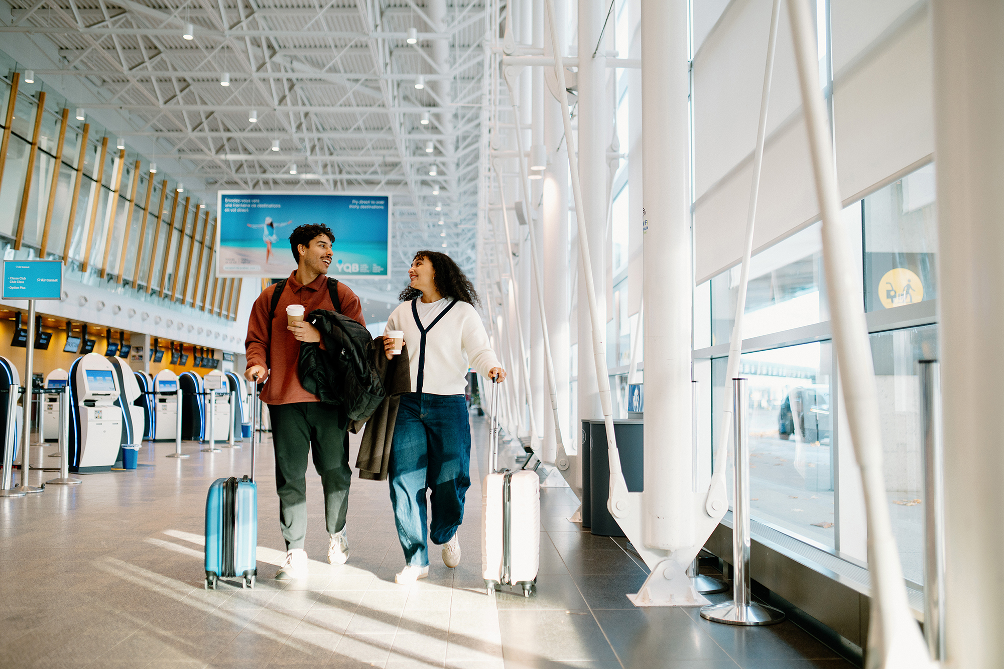 Un homme et une femme traversent le hall d'un aéroport avec leurs valises à roulettes et leurs tasses de café, en se souriant. On aperçoit à l'arrière-plan des bornes d'enregistrement automatiques et de grandes baies vitrées.
