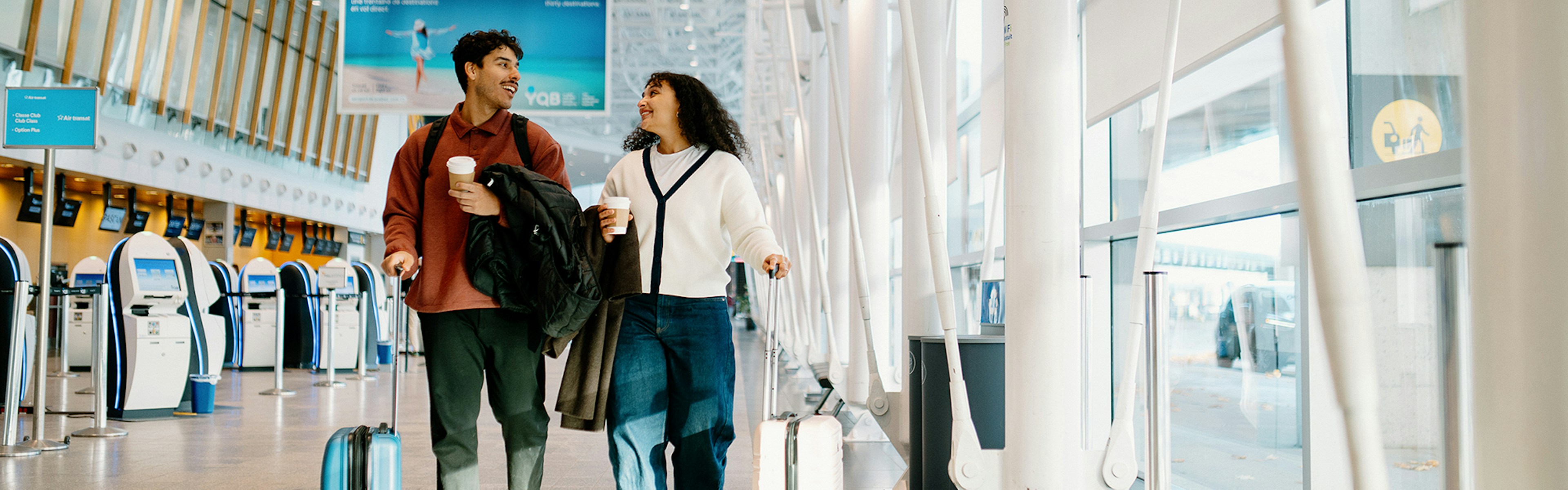 Un homme et une femme traversent le hall d'un aéroport avec leurs valises à roulettes et leurs tasses de café, en se souriant. On aperçoit à l'arrière-plan des bornes d'enregistrement automatiques et de grandes baies vitrées.