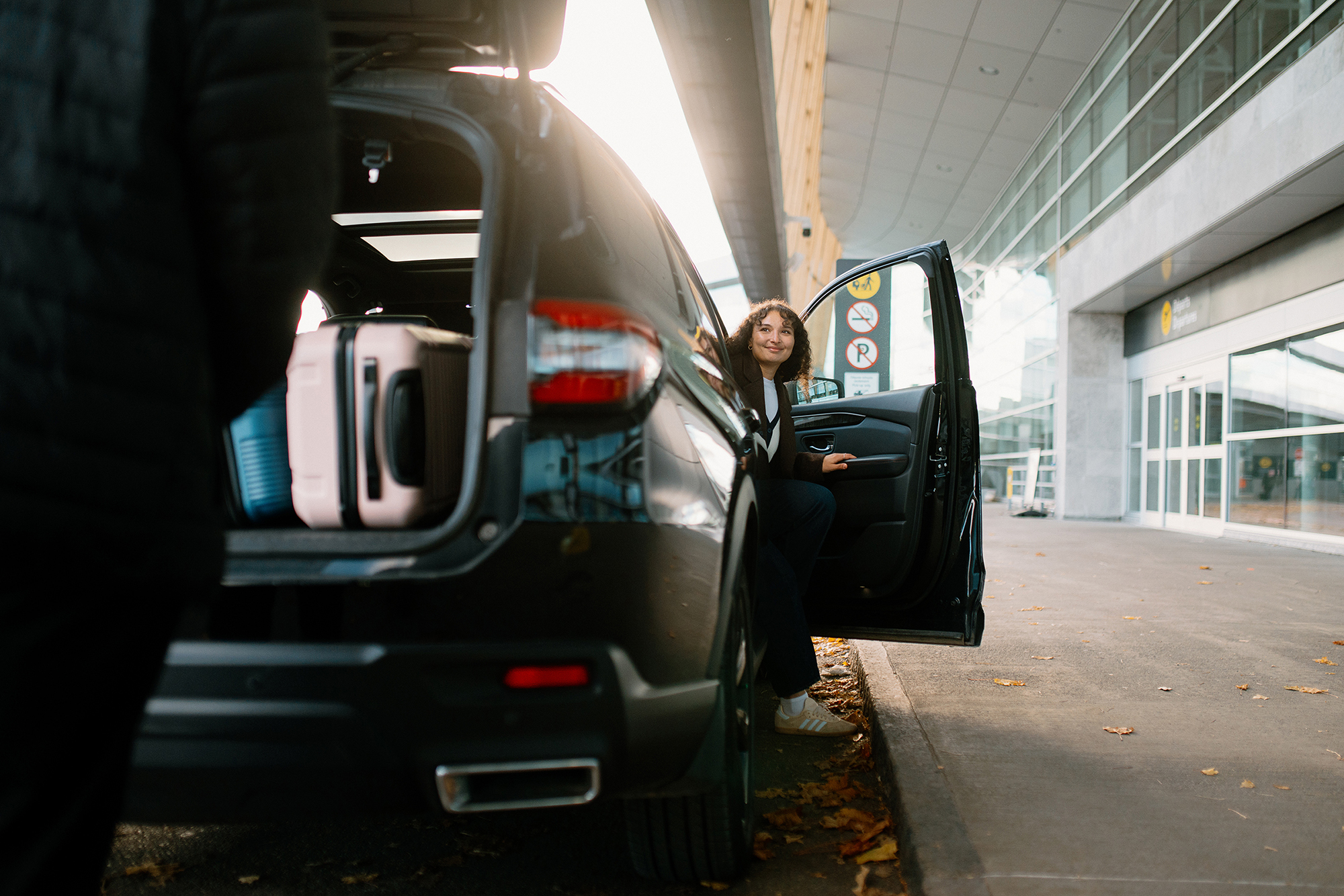Une personne est assise près d'une portière ouverte, devant un bâtiment moderne ; on aperçoit des valises dans le coffre de la voiture. Il semble s'agir d'un aéroport ou d'une zone de transit.