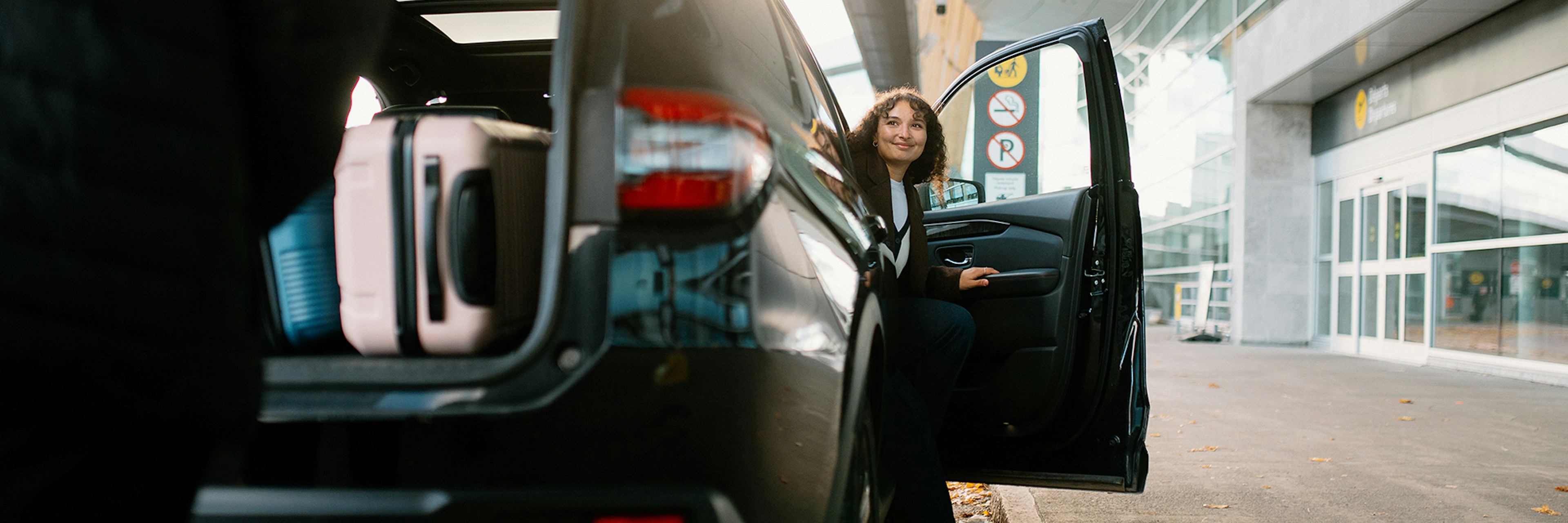 Une personne est assise près d'une portière ouverte, devant un bâtiment moderne ; on aperçoit des valises dans le coffre de la voiture. Il semble s'agir d'un aéroport ou d'une zone de transit.