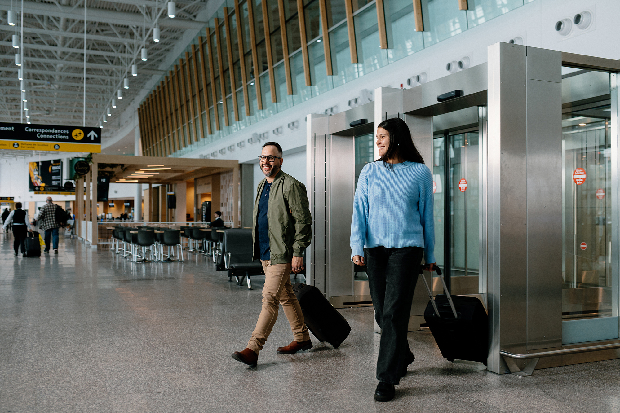 Un homme et une femme, tous deux souriants et tirant chacun une valise, franchissent les portes vitrées d'un terminal d'aéroport moderne aux hauts plafonds et aux grandes baies vitrées.
