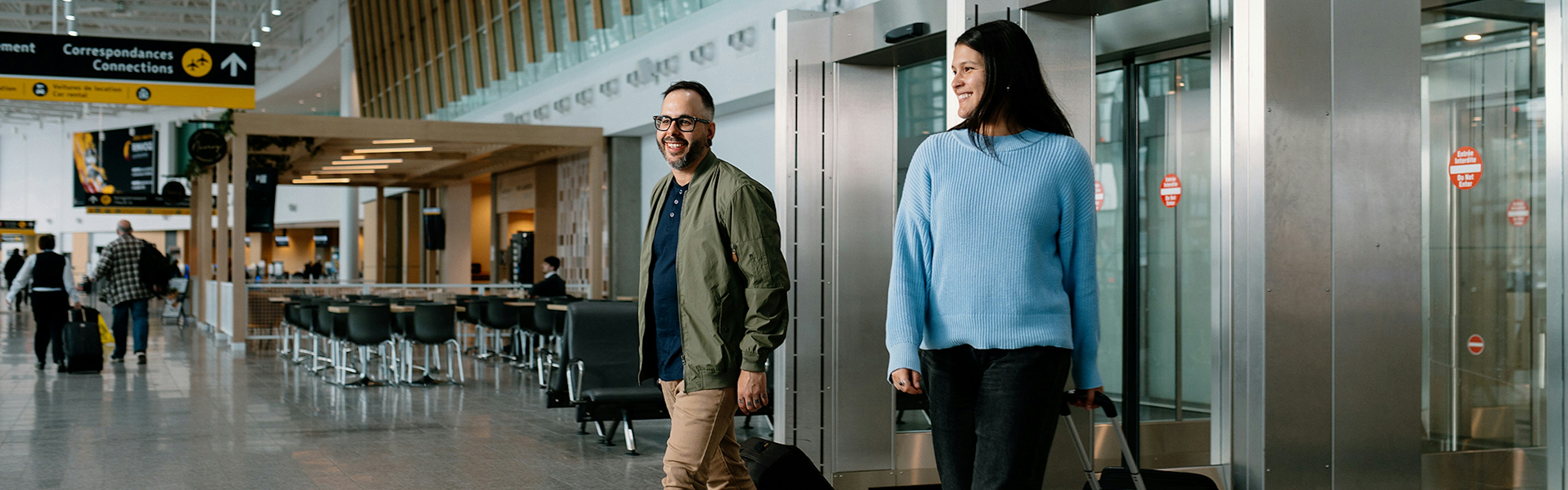 Un homme et une femme, tous deux souriants et tirant chacun une valise, franchissent les portes vitrées d'un terminal d'aéroport moderne aux hauts plafonds et aux grandes baies vitrées.
