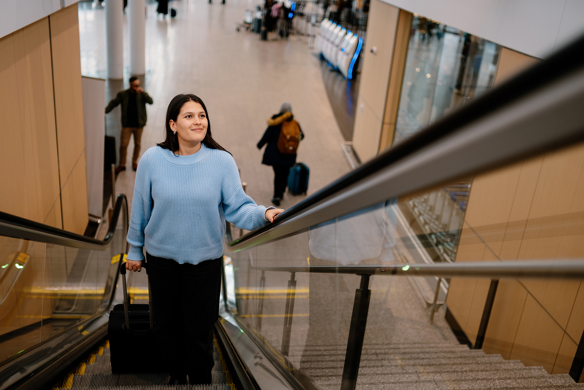 Une femme, une valise à la main, monte un escalator dans un aéroport ; elle porte un pull bleu clair et sourit. On aperçoit d'autres voyageurs et des bagages à l'arrière-plan.