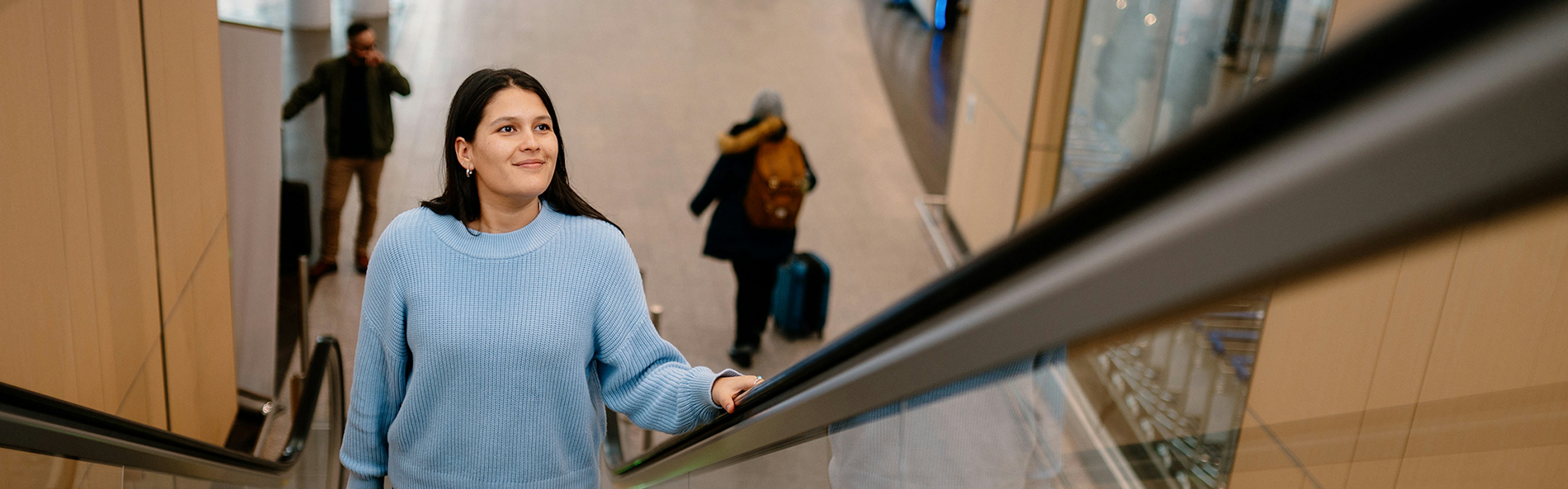 Une femme, une valise à la main, monte un escalator dans un aéroport ; elle porte un pull bleu clair et sourit. On aperçoit d'autres voyageurs et des bagages à l'arrière-plan.