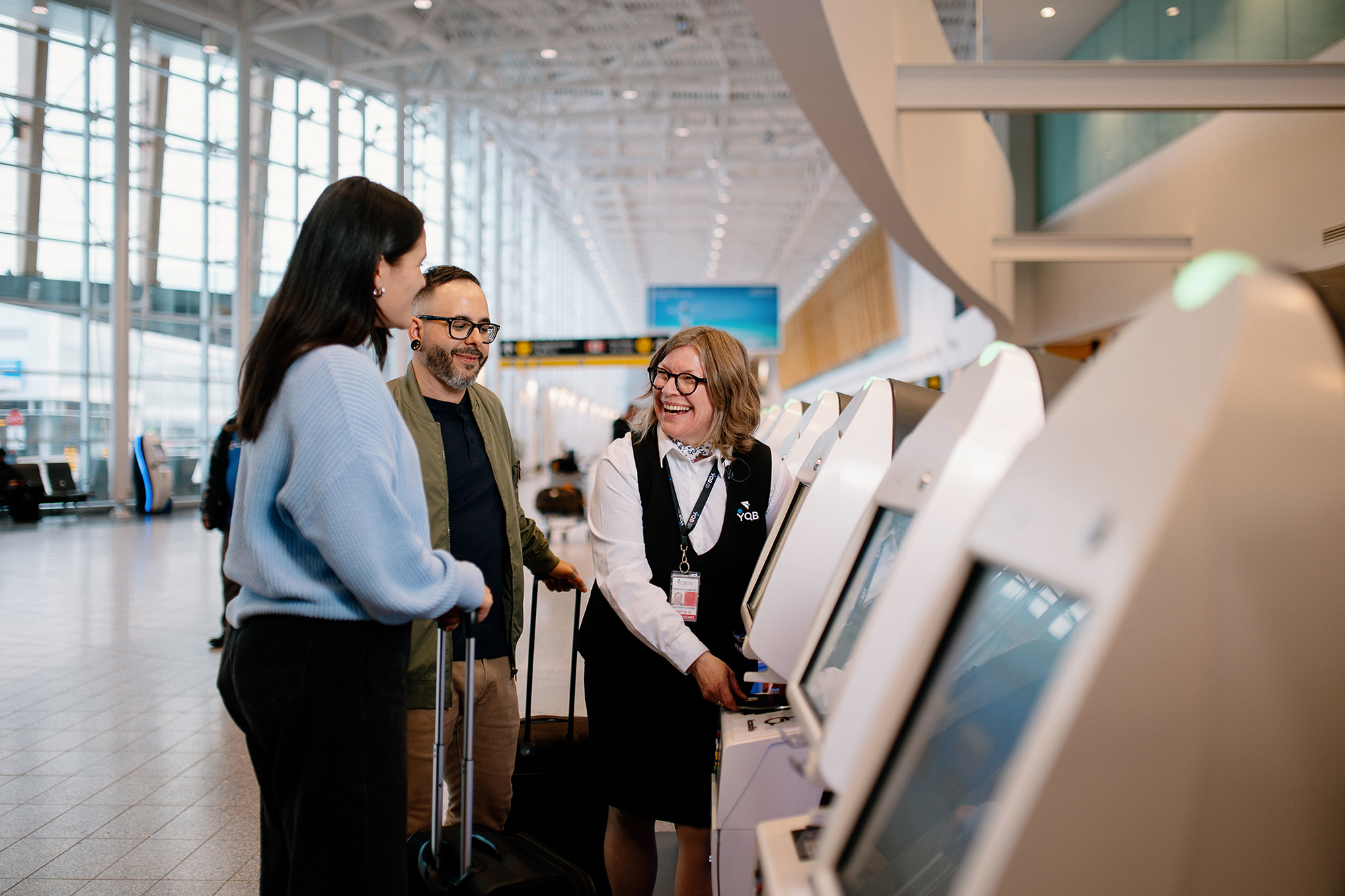 Une femme aide deux voyageurs à utiliser une borne libre-service dans un aéroport. Les voyageurs ont des valises et sourient. Le lieu est lumineux, avec de grandes baies vitrées et une architecture moderne.