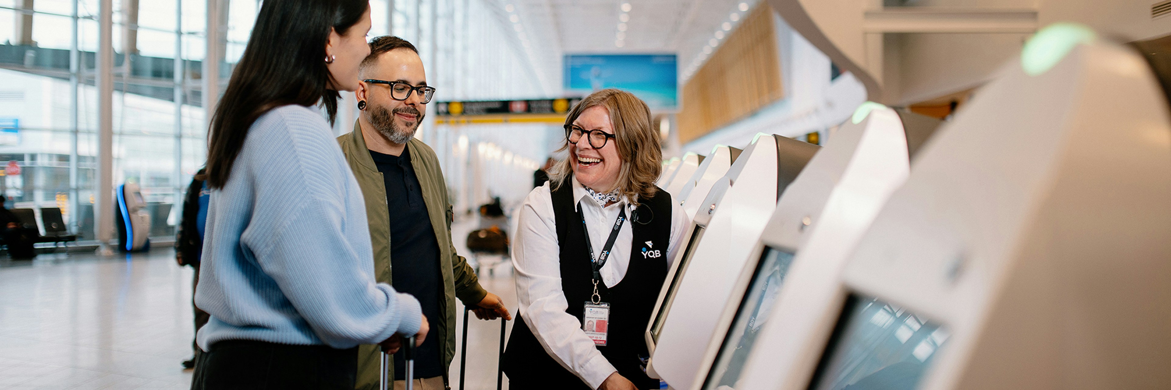 Une femme aide deux voyageurs à utiliser une borne libre-service dans un aéroport. Les voyageurs ont des valises et sourient. Le lieu est lumineux, avec de grandes baies vitrées et une architecture moderne.