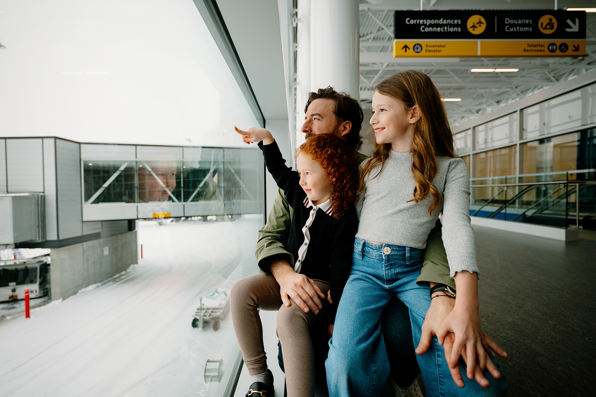 Un homme est assis avec deux jeunes filles près d'une fenêtre de l'aéroport. L'une des filles montre du doigt la piste enneigée à l'extérieur tandis que l'autre sourit ; on aperçoit au-dessus d'elles des panneaux jaunes indiquant les guichets du courrier et des douanes.