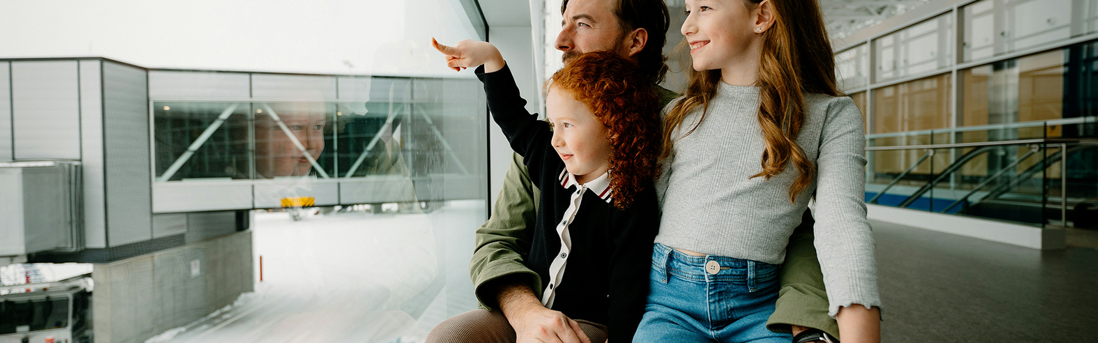 Un homme est assis avec deux jeunes filles près d'une fenêtre de l'aéroport. L'une des filles montre du doigt la piste enneigée à l'extérieur tandis que l'autre sourit ; on aperçoit au-dessus d'elles des panneaux jaunes indiquant les guichets du courrier et des douanes.