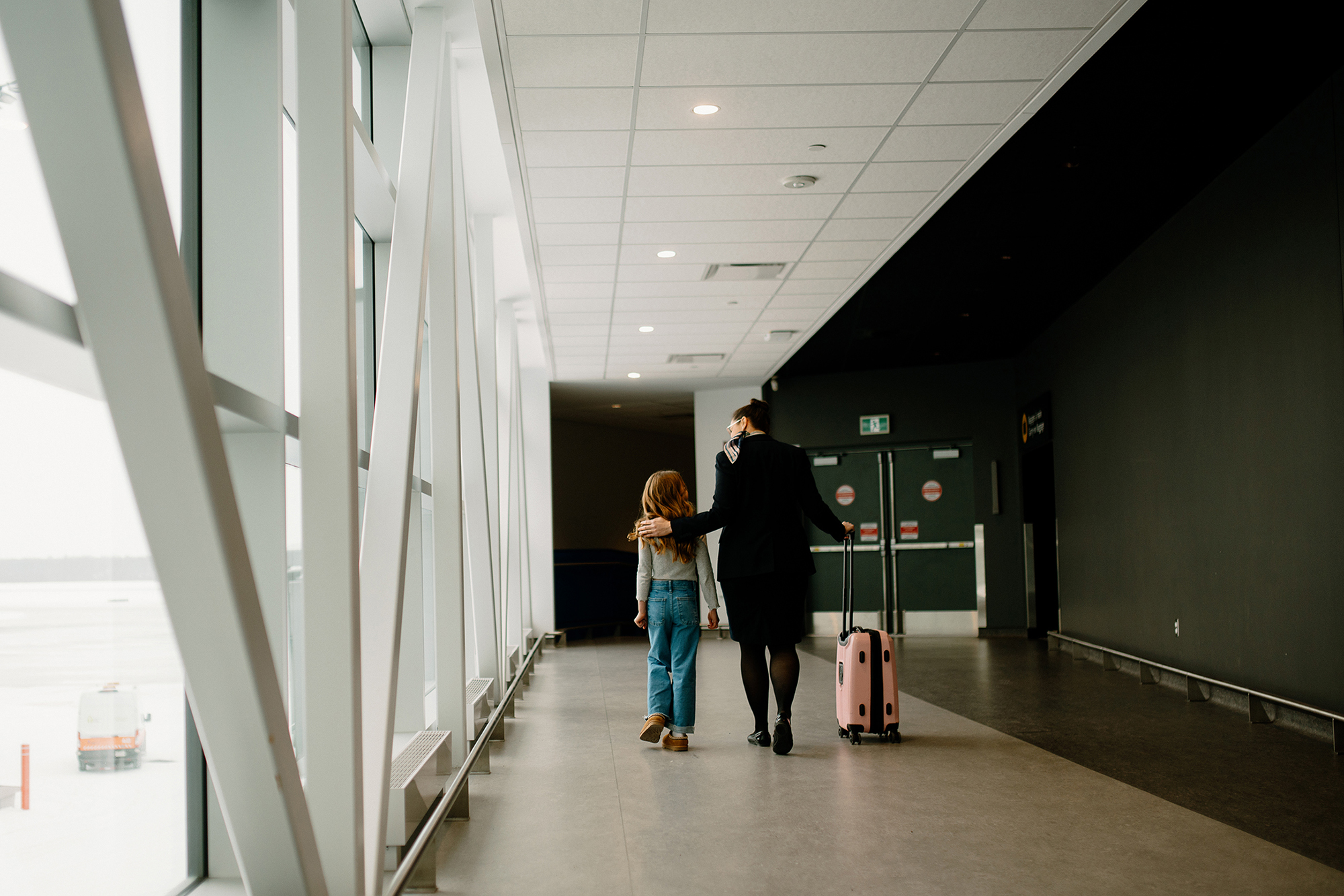 Un enfant et un adulte marchent côte à côte dans un couloir de l'aéroport. L'adulte a son bras autour des épaules de l'enfant et tire une valise rose. De grandes baies vitrées bordent l'un des côtés, laissant entrer la lumière naturelle.