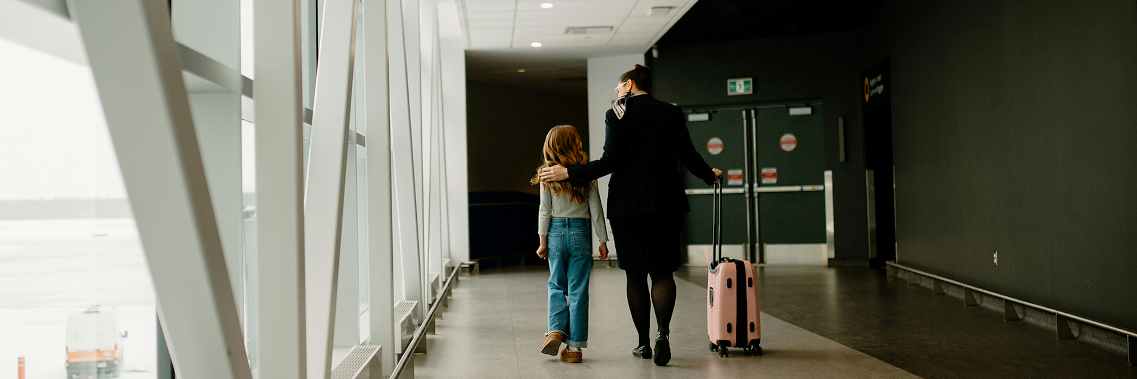 Un enfant et un adulte marchent côte à côte dans un couloir de l'aéroport. L'adulte a son bras autour des épaules de l'enfant et tire une valise rose. De grandes baies vitrées bordent l'un des côtés, laissant entrer la lumière naturelle.