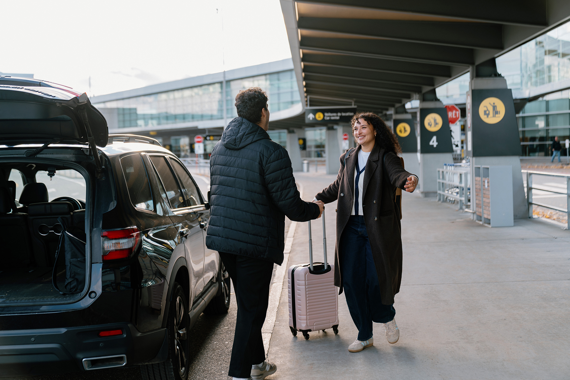 Une personne munie d'une valise salue une autre personne près d'une voiture, à l'extérieur du terminal de l'aéroport ; toutes deux sourient et se tendent la main.