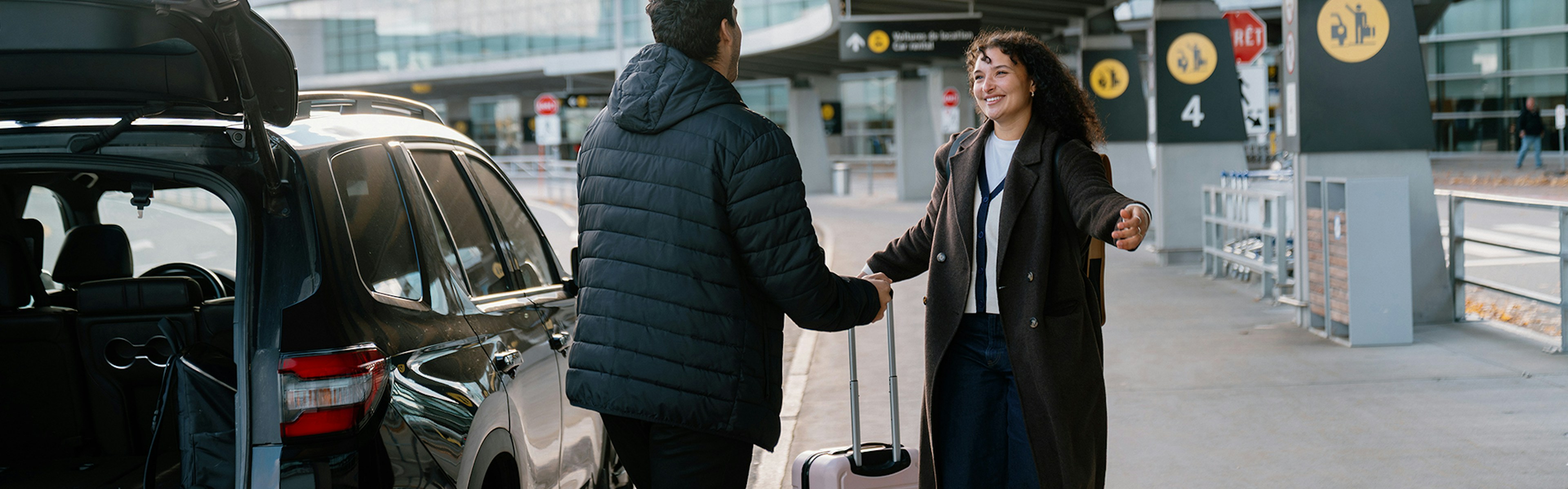 Une personne munie d'une valise salue une autre personne près d'une voiture, à l'extérieur du terminal de l'aéroport ; toutes deux sourient et se tendent la main.