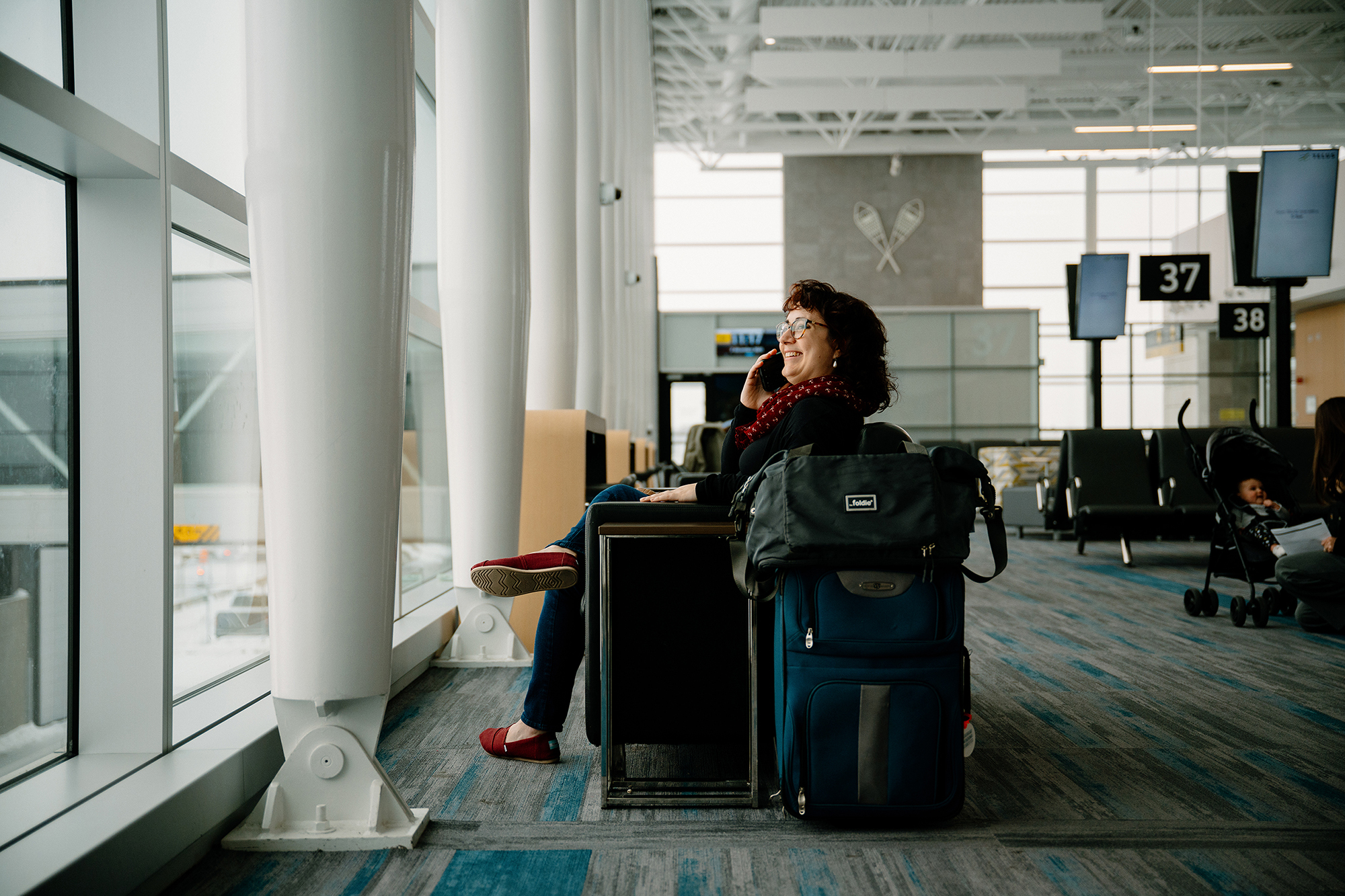 Woman sitting by a window at an airport gate, smiling while talking on her phone. She has a suitcase and backpack next to her. Empty seats and boarding gates are visible in the background.
