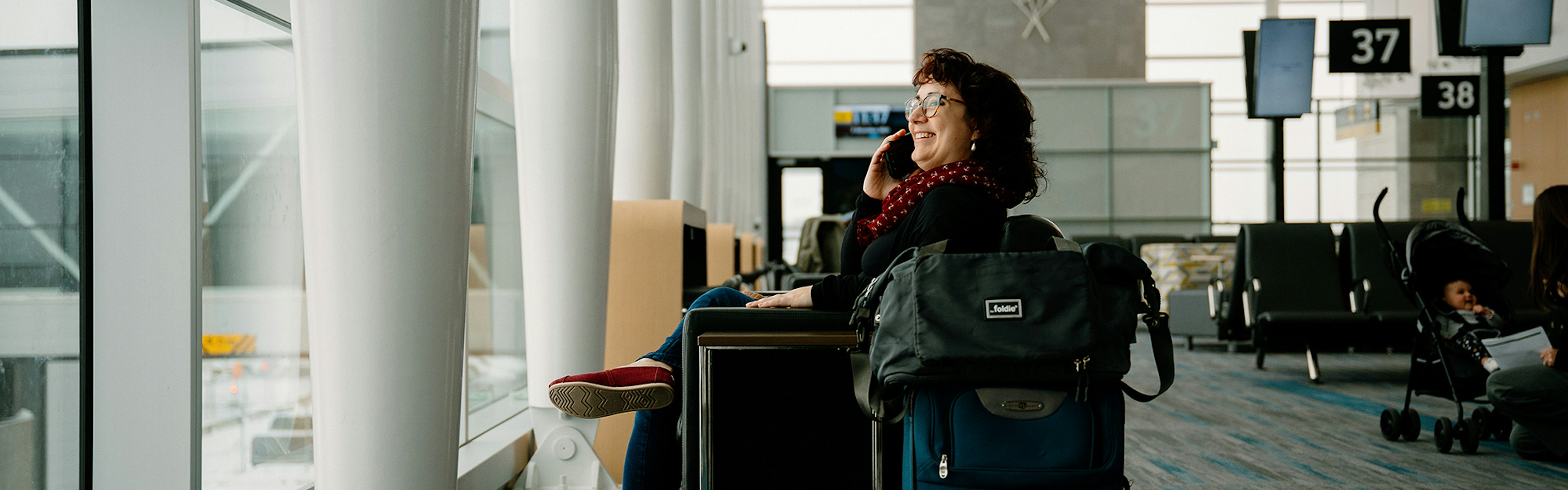 Woman sitting by a window at an airport gate, smiling while talking on her phone. She has a suitcase and backpack next to her. Empty seats and boarding gates are visible in the background.