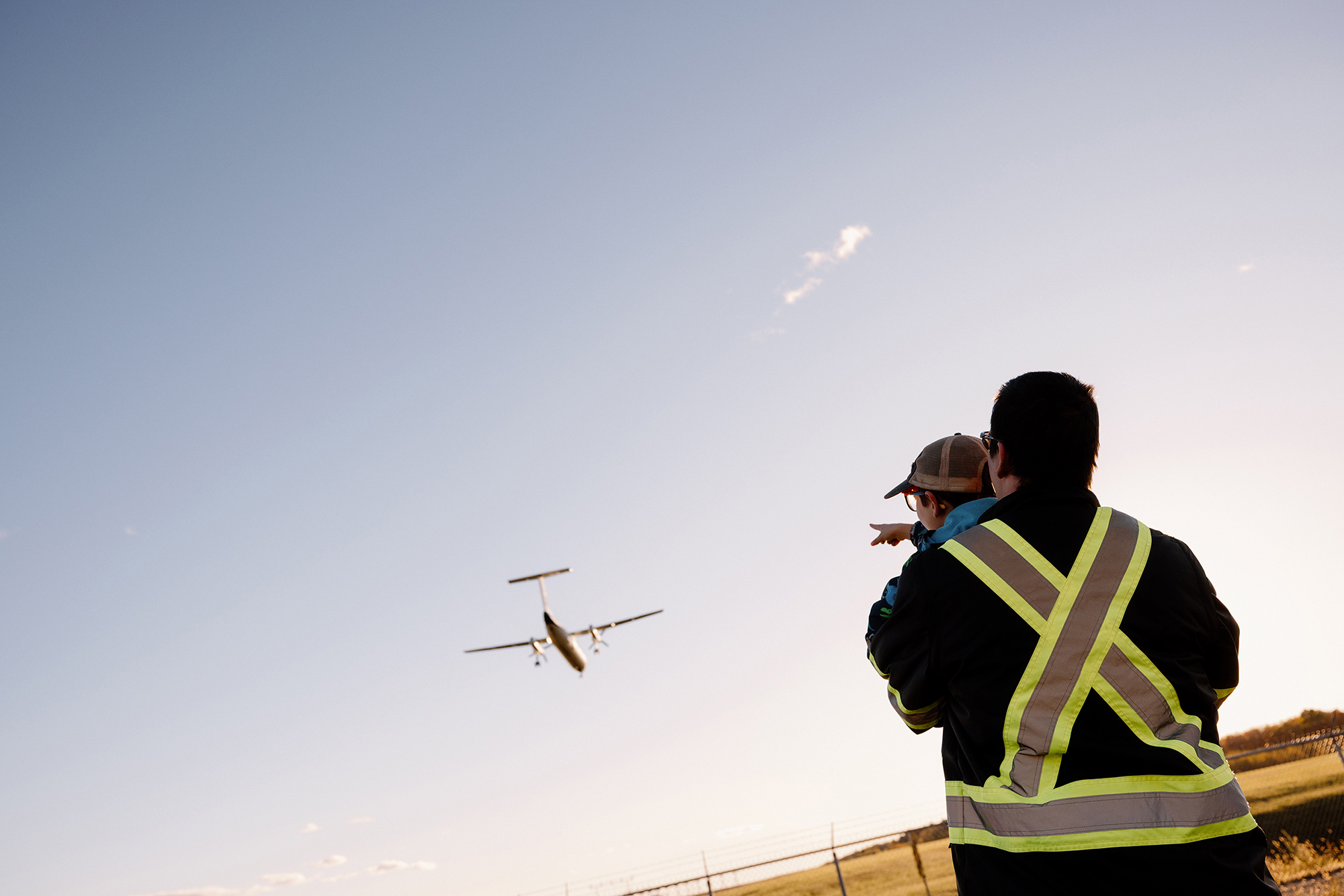 Un adulte vêtu d'un gilet réfléchissant tient un enfant dans ses bras tandis qu'ils regardent tous les deux un petit avion volant à basse altitude au coucher du soleil.
