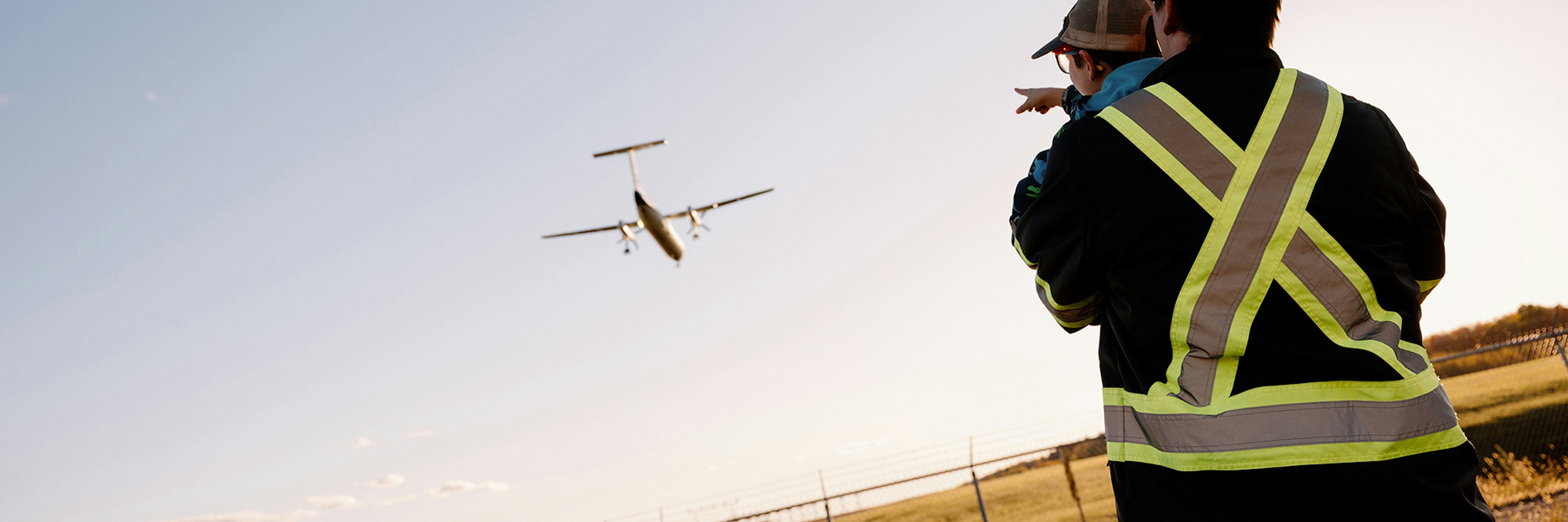 Un adulte vêtu d'un gilet réfléchissant tient un enfant dans ses bras tandis qu'ils regardent tous les deux un petit avion volant à basse altitude au coucher du soleil.