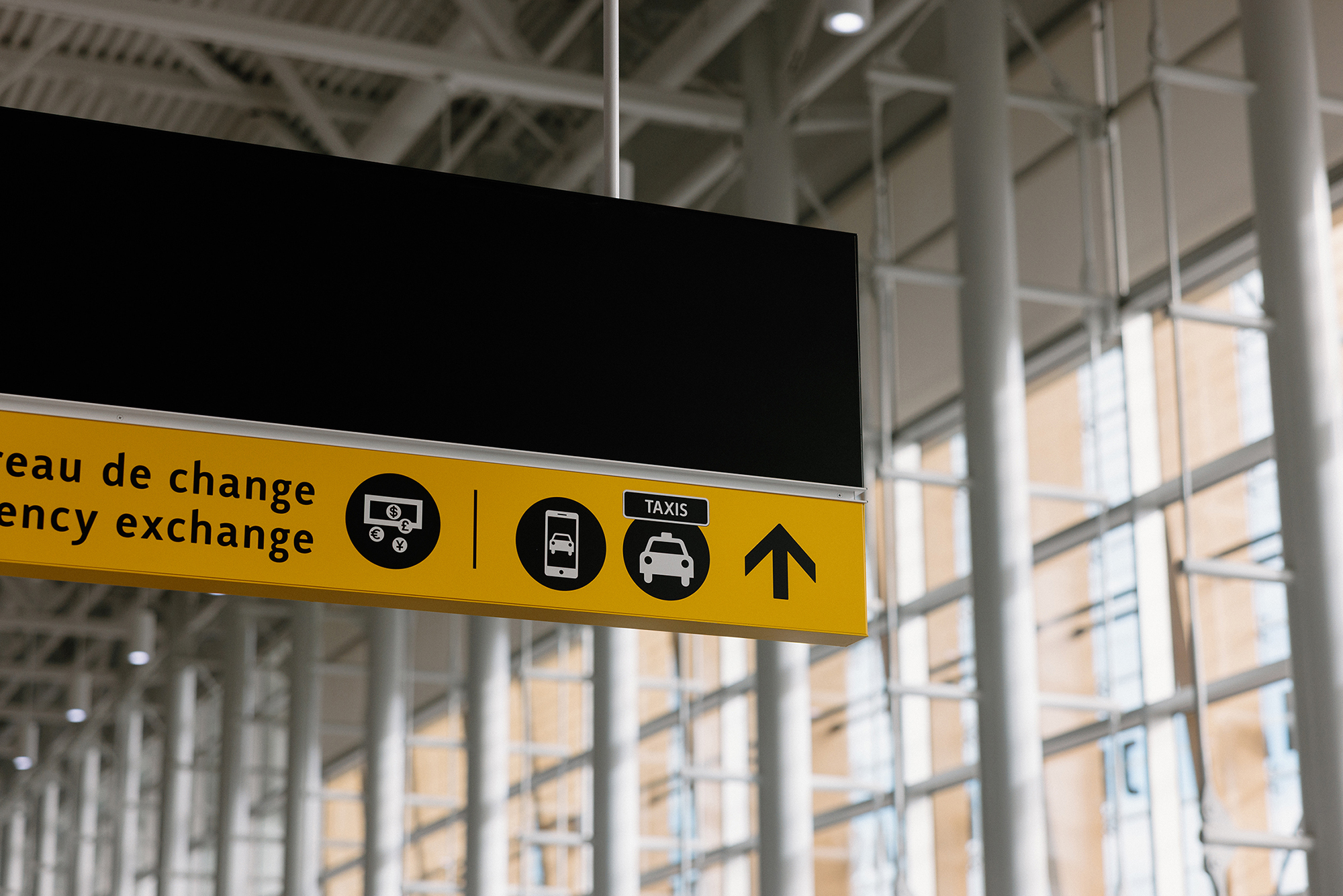 An airport sign featuring icons representing currency exchange, buses, taxis, and an arrow indicating the direction to follow. The background shows large floor-to-ceiling windows and structural columns inside the terminal.