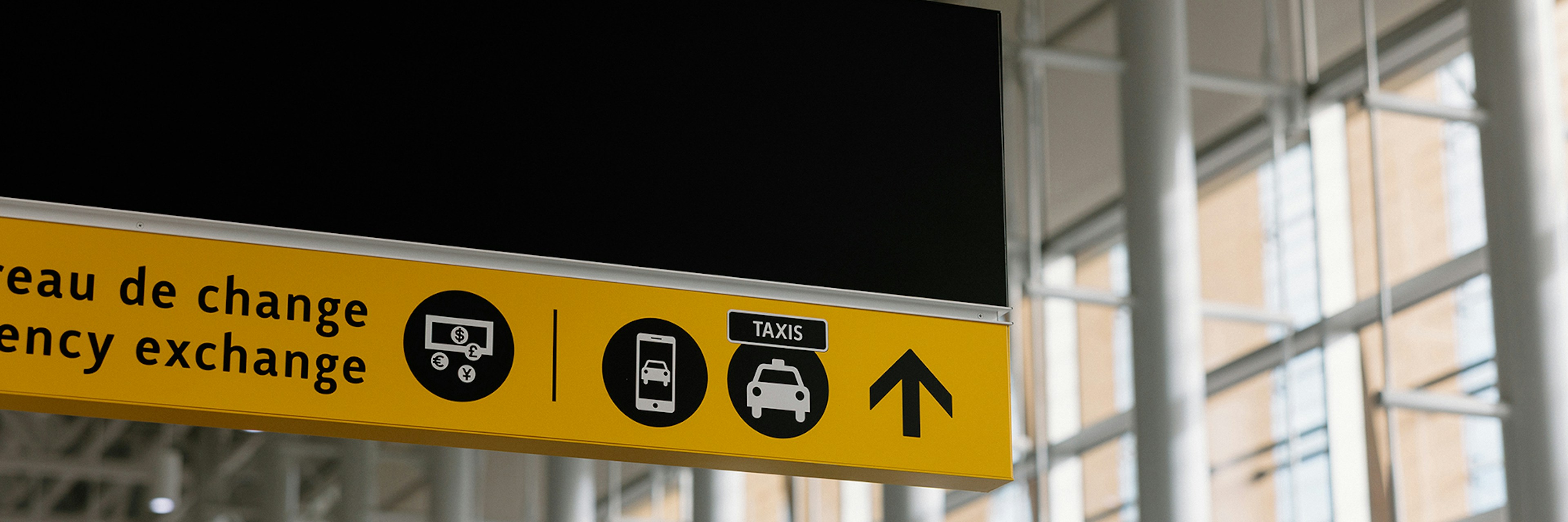 An airport sign featuring icons representing currency exchange, buses, taxis, and an arrow indicating the direction to follow. The background shows large floor-to-ceiling windows and structural columns inside the terminal.