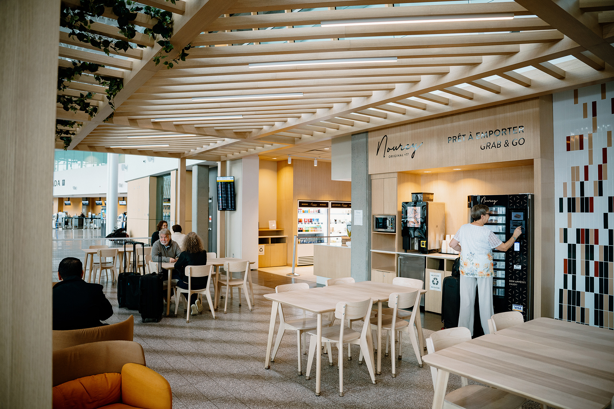 Un café d'aéroport moderne, avec des tables et des chaises en bois, des distributeurs automatiques et quelques personnes assises ou en train de commander à manger. De grandes baies vitrées laissent entrer la lumière naturelle, et on aperçoit des panneaux d'affichage numériques des vols en arrière-plan.