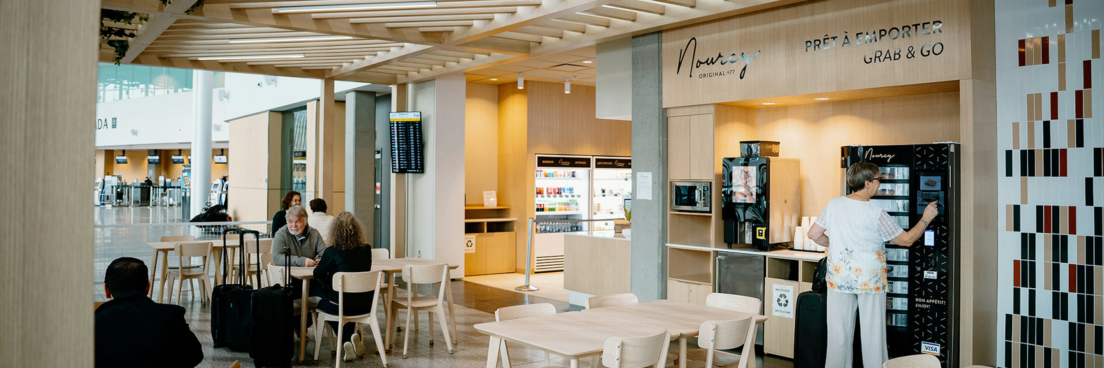 Un café d'aéroport moderne, avec des tables et des chaises en bois, des distributeurs automatiques et quelques personnes assises ou en train de commander à manger. De grandes baies vitrées laissent entrer la lumière naturelle, et on aperçoit des panneaux d'affichage numériques des vols en arrière-plan.