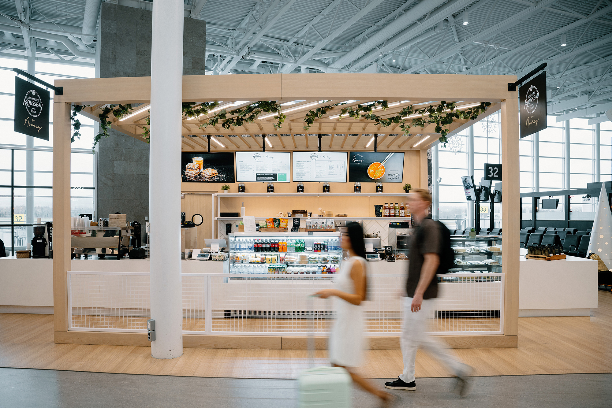 Un café d'aéroport moderne, avec des touches de bois, des panneaux de menu numériques et un présentoir proposant des en-cas et des boissons. Un homme et une femme, chargés de bagages, passent devant, tandis que la porte d'embarquement n° 32 est visible à l'arrière-plan.