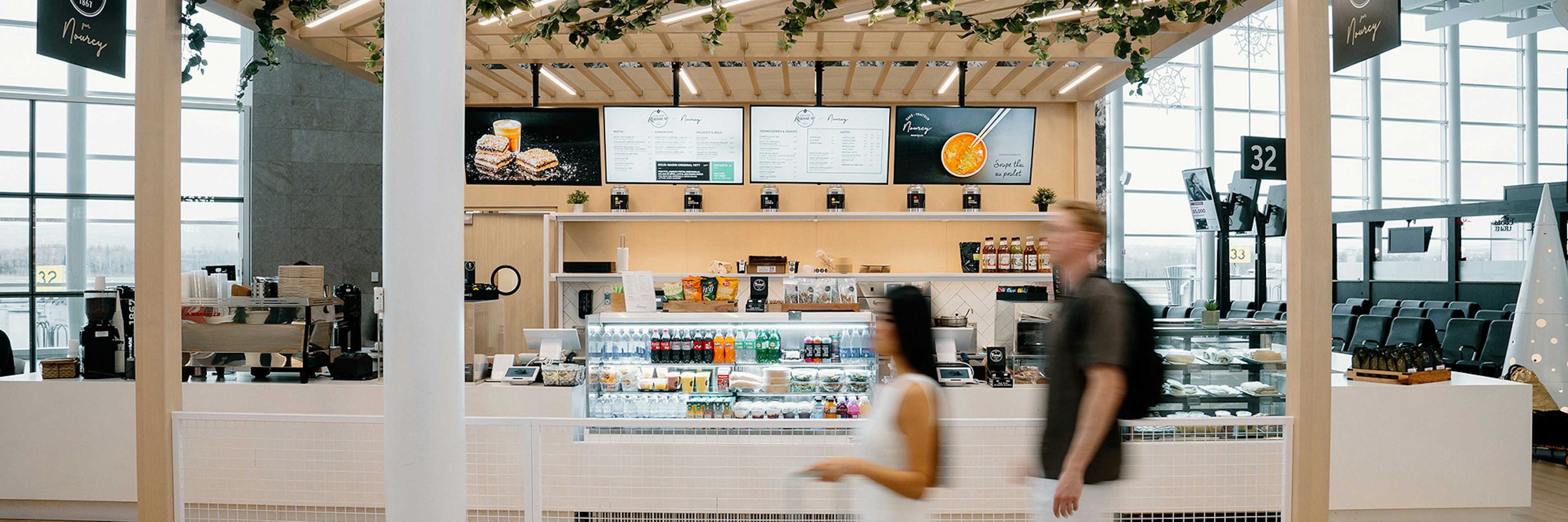 Un café d'aéroport moderne, avec des touches de bois, des panneaux de menu numériques et un présentoir proposant des en-cas et des boissons. Un homme et une femme, chargés de bagages, passent devant, tandis que la porte d'embarquement n° 32 est visible à l'arrière-plan.