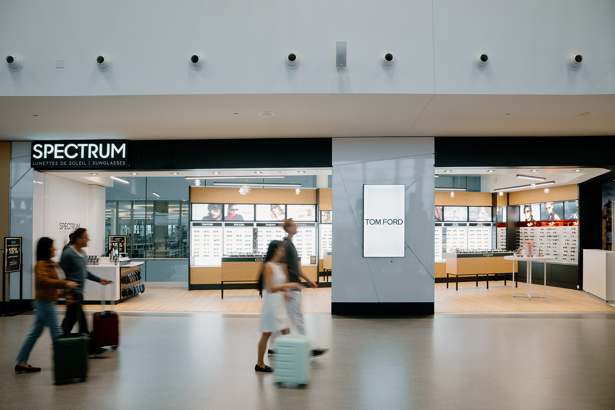 Trois personnes, chargées de bagages, passent devant une boutique moderne de lunettes de soleil appelée « Spectrum » dans un aéroport. La boutique présente des vitrines aux couleurs vives et une publicité pour Tom Ford accrochée au mur au centre.