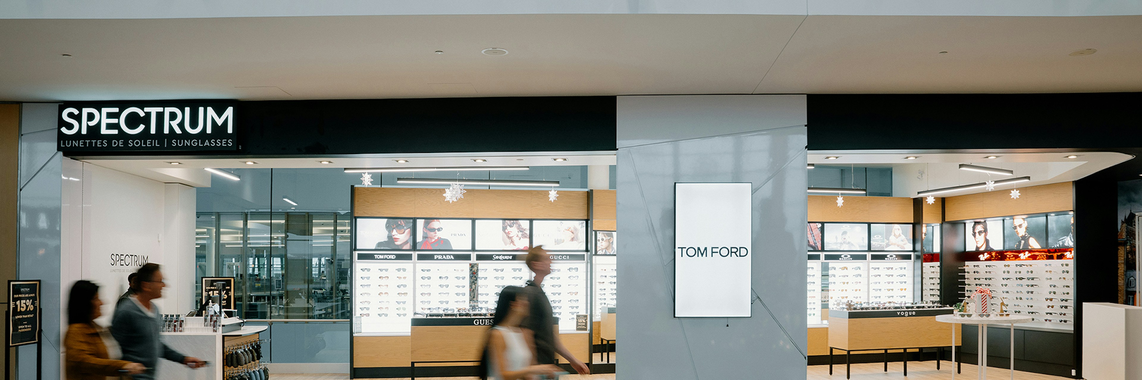 Trois personnes, chargées de bagages, passent devant une boutique moderne de lunettes de soleil appelée « Spectrum » dans un aéroport. La boutique présente des vitrines aux couleurs vives et une publicité pour Tom Ford accrochée au mur au centre.