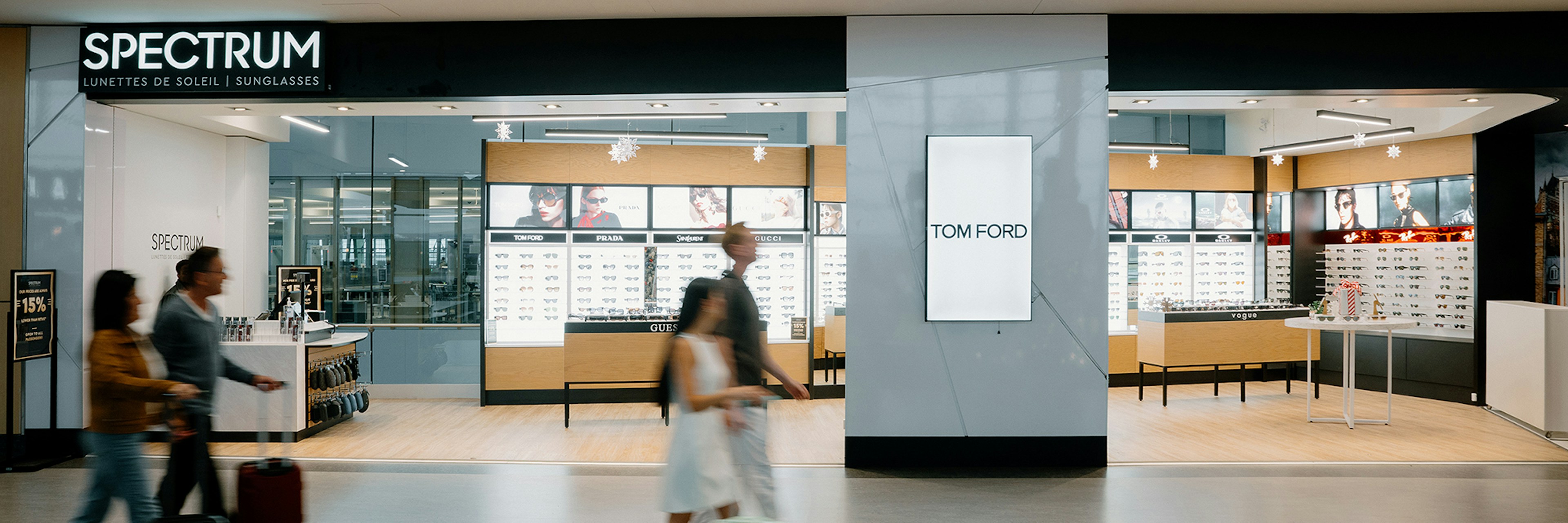 People with luggage walk past a modern airport store called Spectrum, which sells sunglasses. The store has displays of eyewear and a Tom Ford advertisement on a wall. The setting is clean and brightly lit.