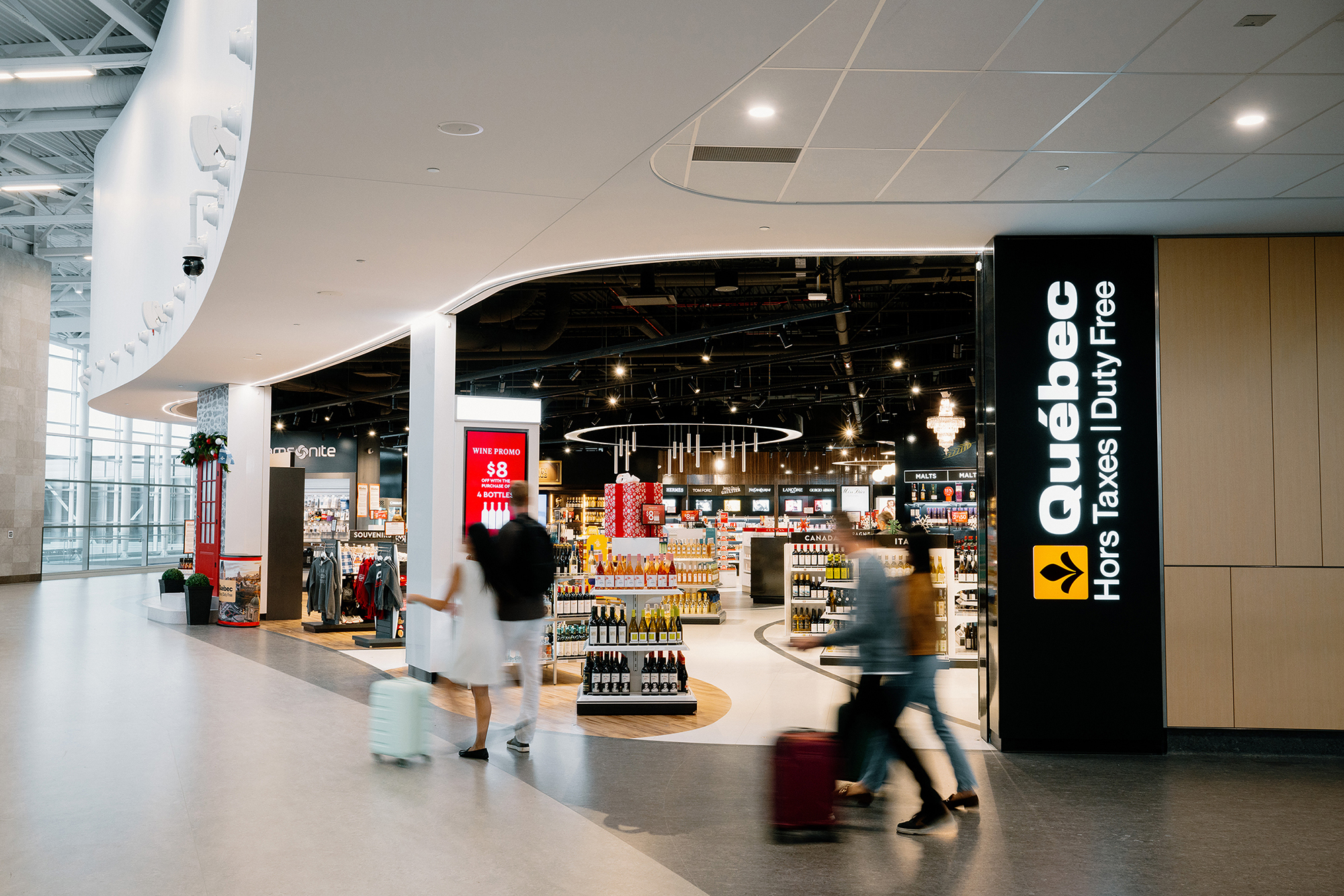 Des voyageurs munis de valises passent devant une boutique « Québec Duty Free » brillamment éclairée dans un aéroport, où l'on aperçoit des rayons remplis de produits et une signalétique claire, en français et en anglais.