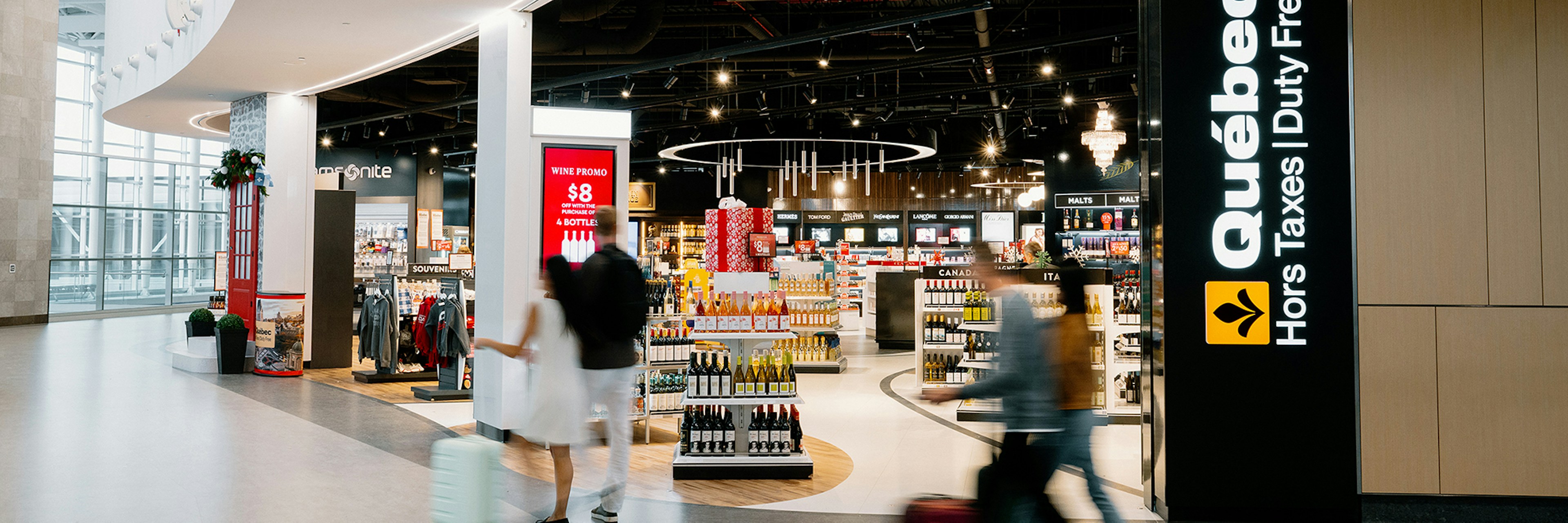 Des voyageurs munis de valises passent devant une boutique « Québec Duty Free » brillamment éclairée dans un aéroport, où l'on aperçoit des rayons remplis de produits et une signalétique claire, en français et en anglais.