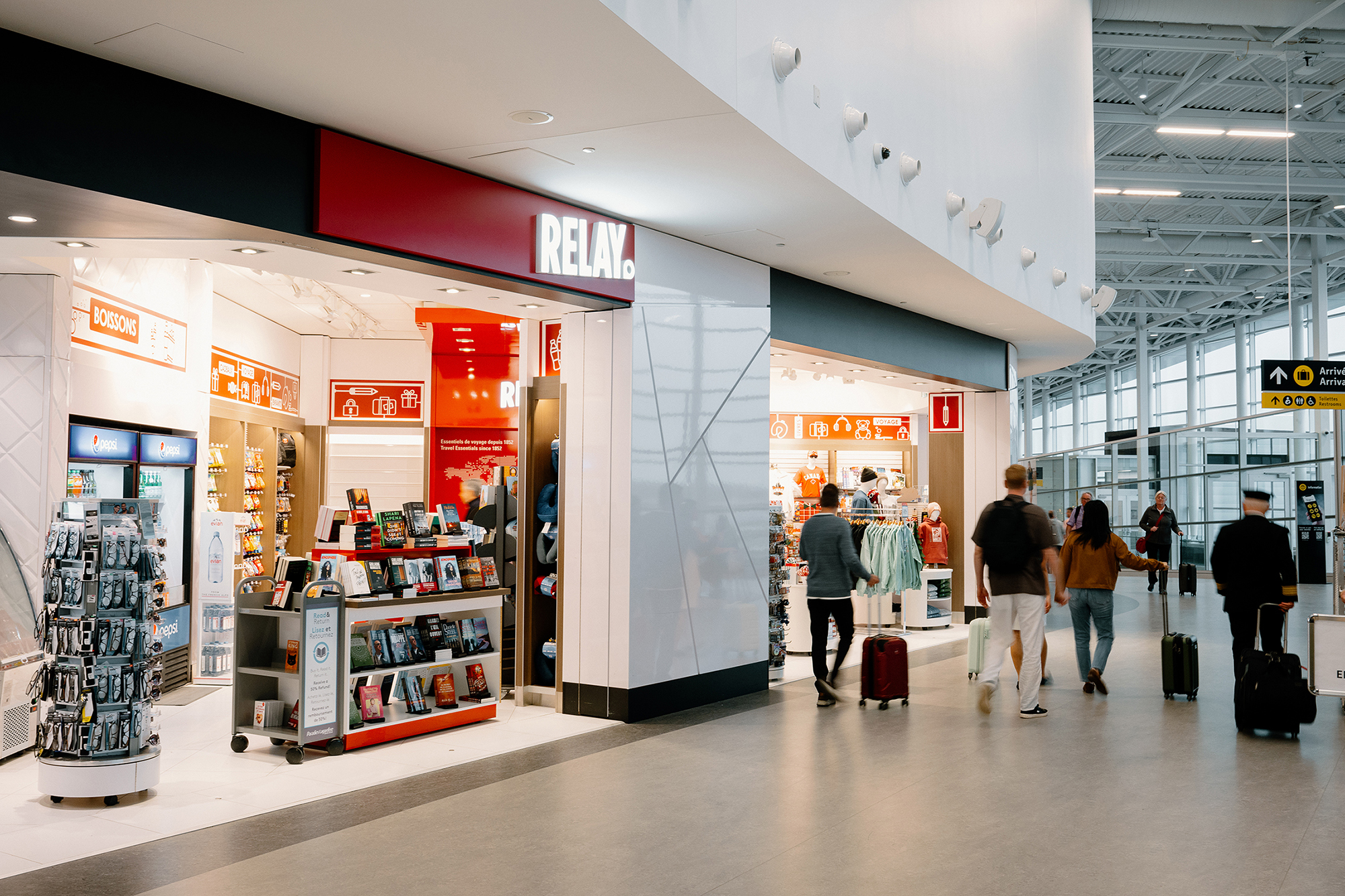 Des passants passent devant une librairie RELAY et une boutique de vêtements situées dans un terminal d'aéroport moderne, où sont exposés des livres, des magazines et des accessoires de voyage.