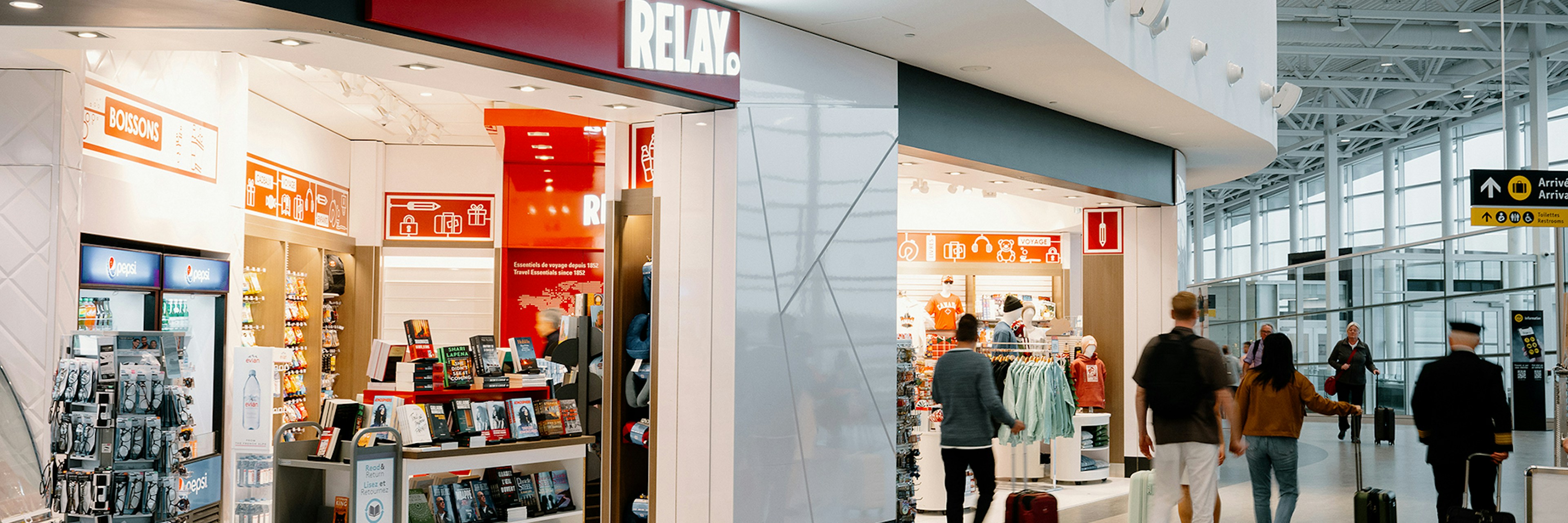 Des passants passent devant une librairie RELAY et une boutique de vêtements situées dans un terminal d'aéroport moderne, où sont exposés des livres, des magazines et des accessoires de voyage.