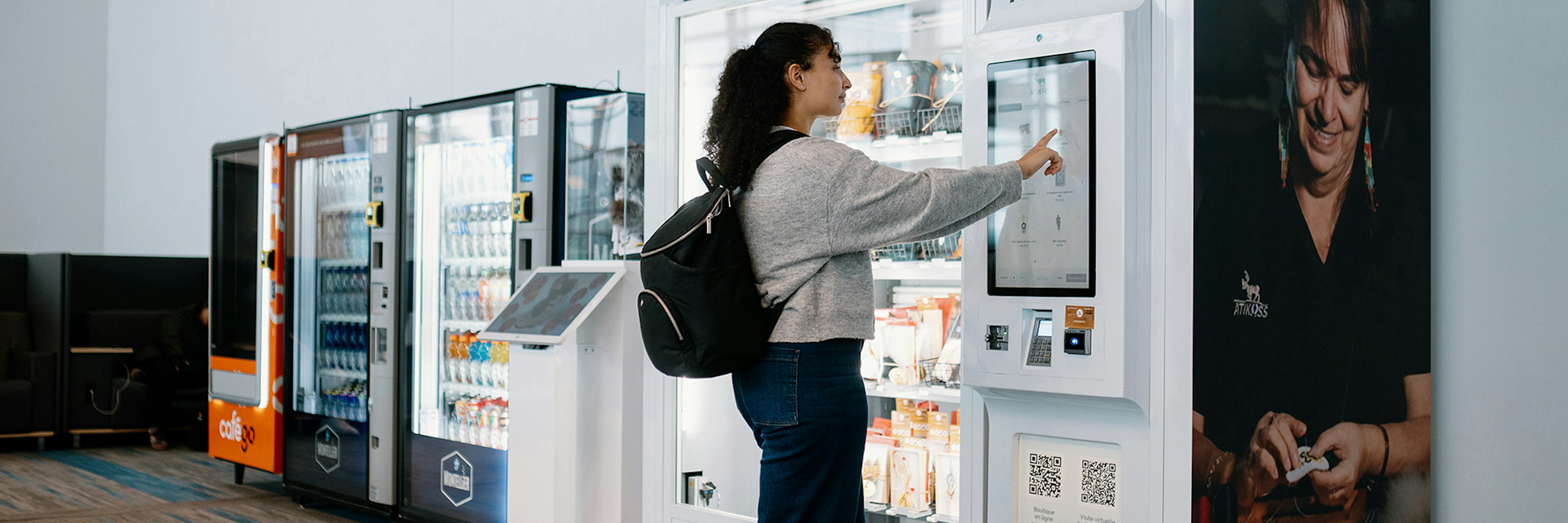 Une femme munie d'un sac à dos utilise l'écran tactile d'un distributeur automatique moderne dans un espace intérieur bien éclairé, avec d'autres distributeurs automatiques visibles à l'arrière-plan.