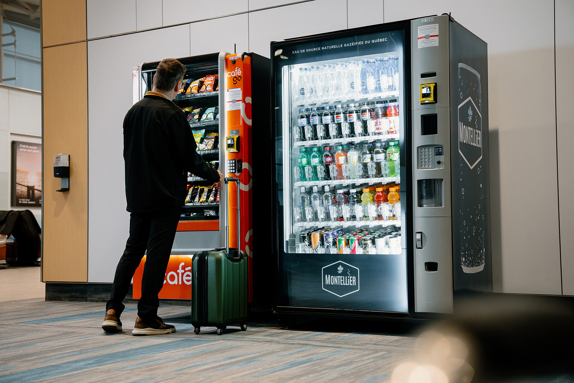 Une personne munie d'une valise utilise un distributeur automatique dans un lieu public couvert, à côté duquel se trouve un autre distributeur proposant des en-cas.