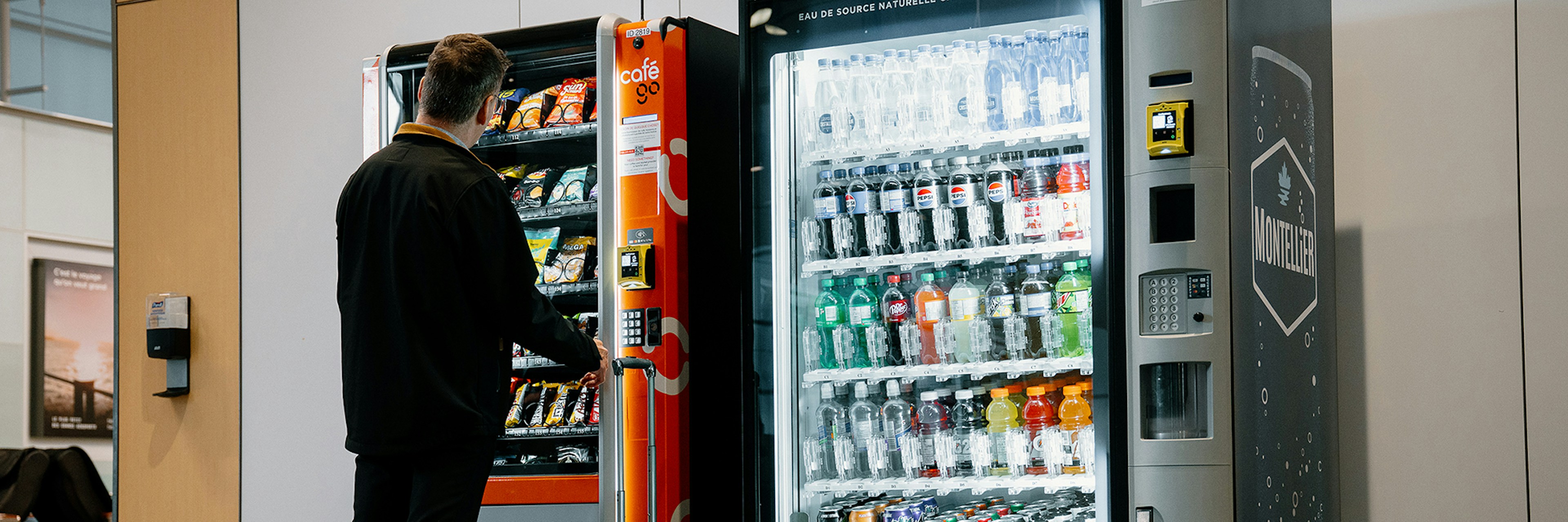 Une personne munie d'une valise utilise un distributeur automatique dans un lieu public couvert, à côté duquel se trouve un autre distributeur proposant des en-cas.