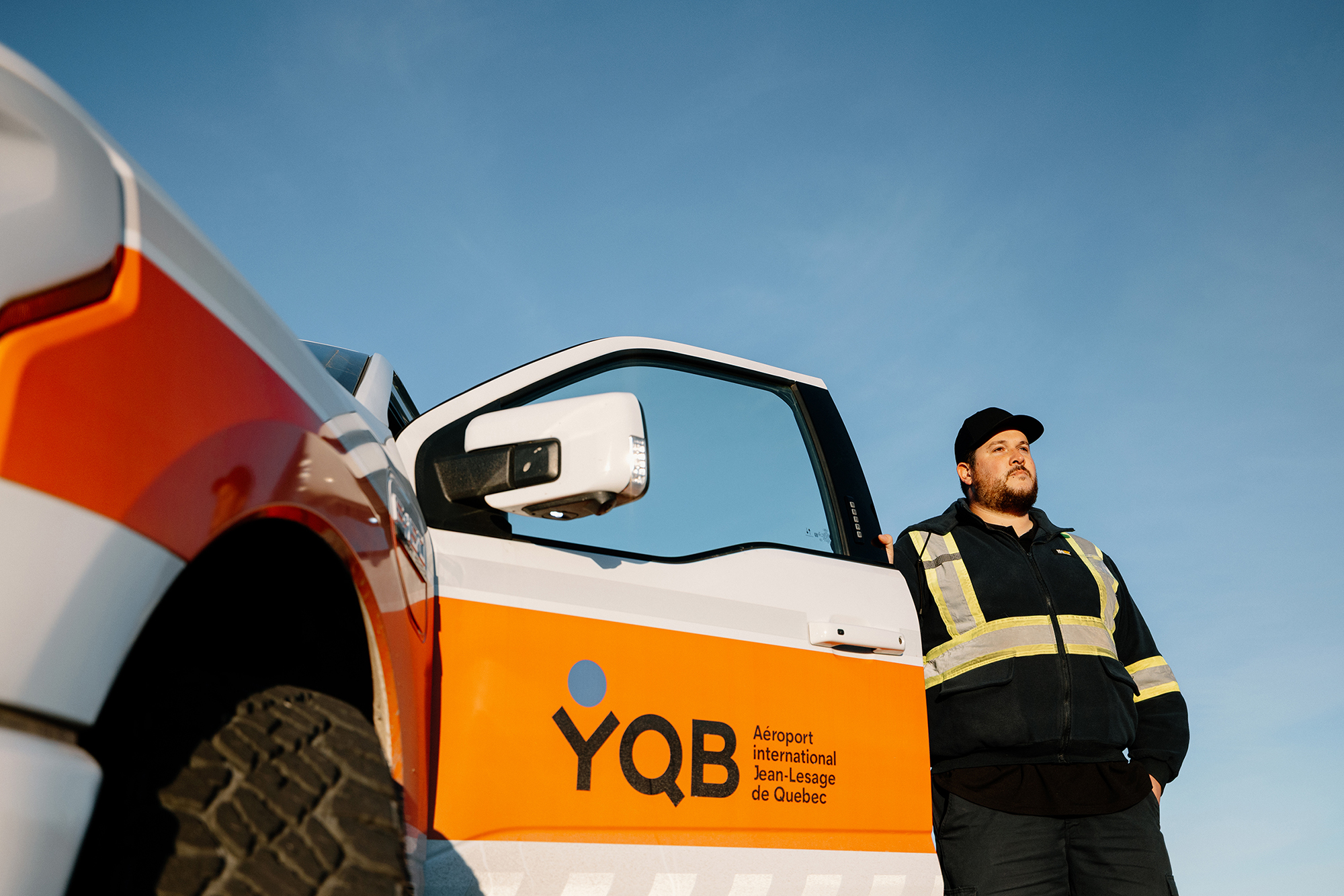 Under a clear blue sky, a man wearing a safety vest stands next to a vehicle bearing the inscription “YQB Jean-Lesage International Airport, Quebec City.”.